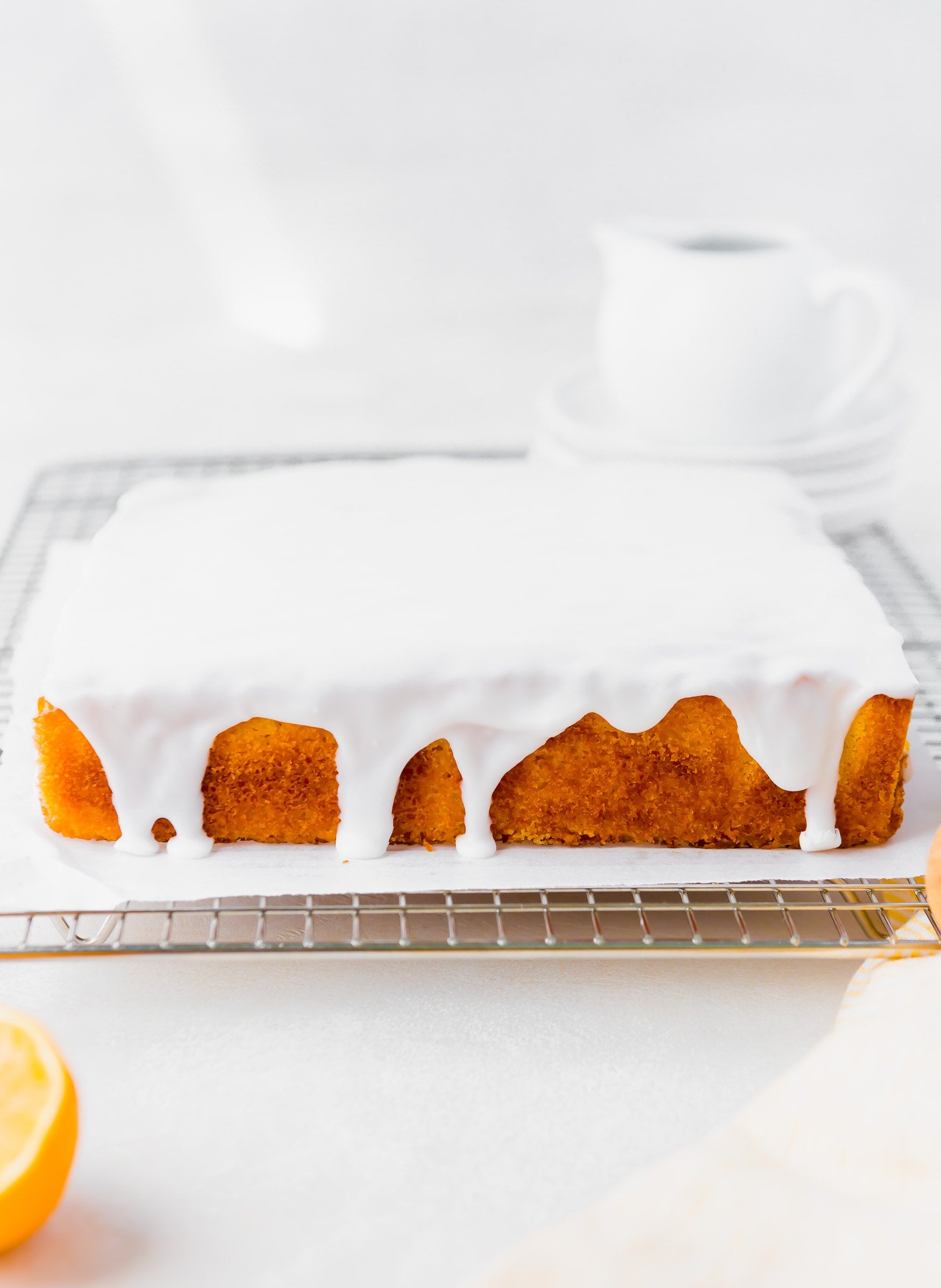 a photo of a square lemon cake covered in a white lemon glaze dripping down the sides. the cake is sitting on a piece of parchment paper on top of a gold cooling rack.