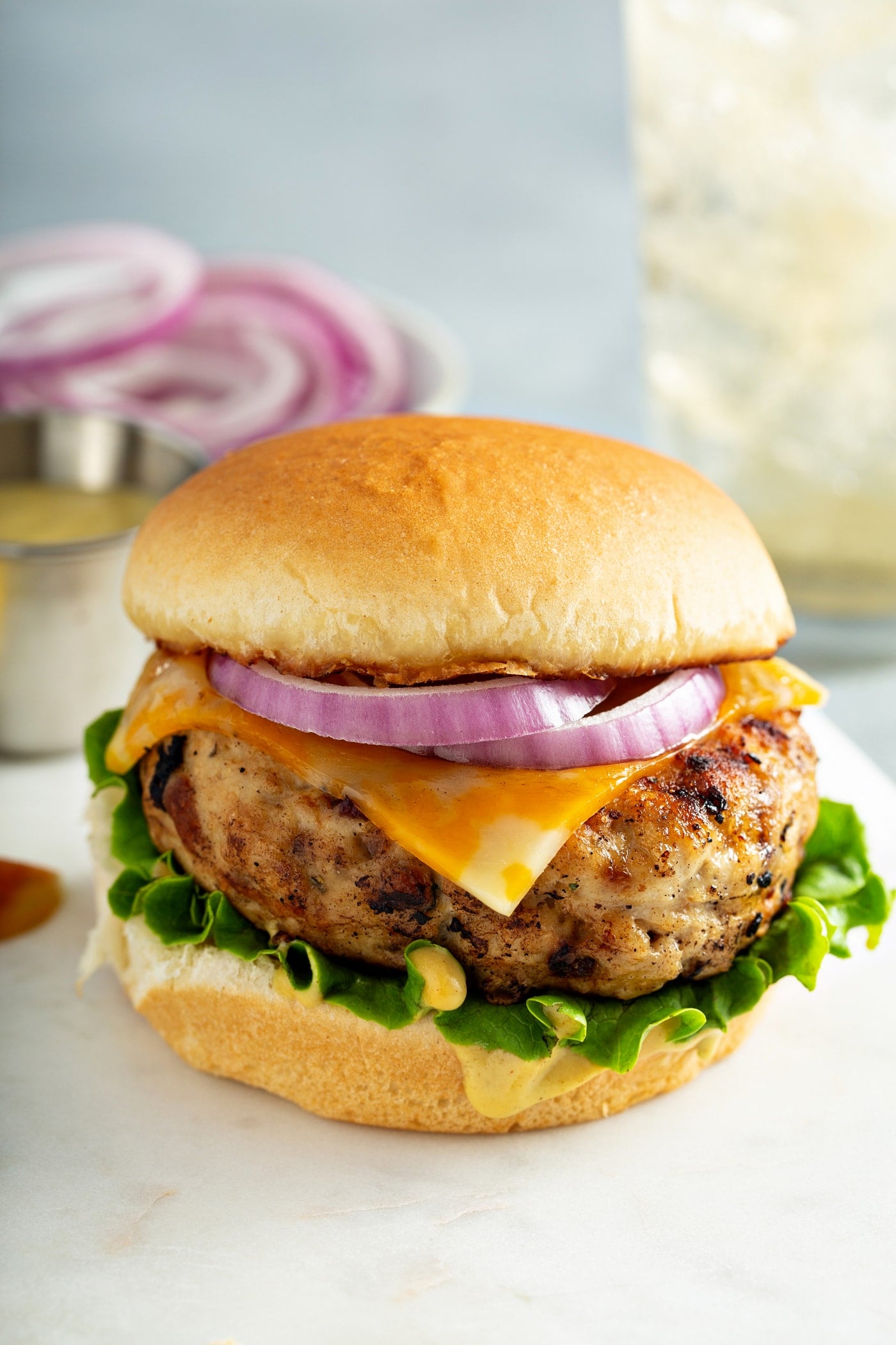 A maple mustard chicken burger on a bun with lettuce, cheese and onions. There is a plate of sliced red onions in the background.