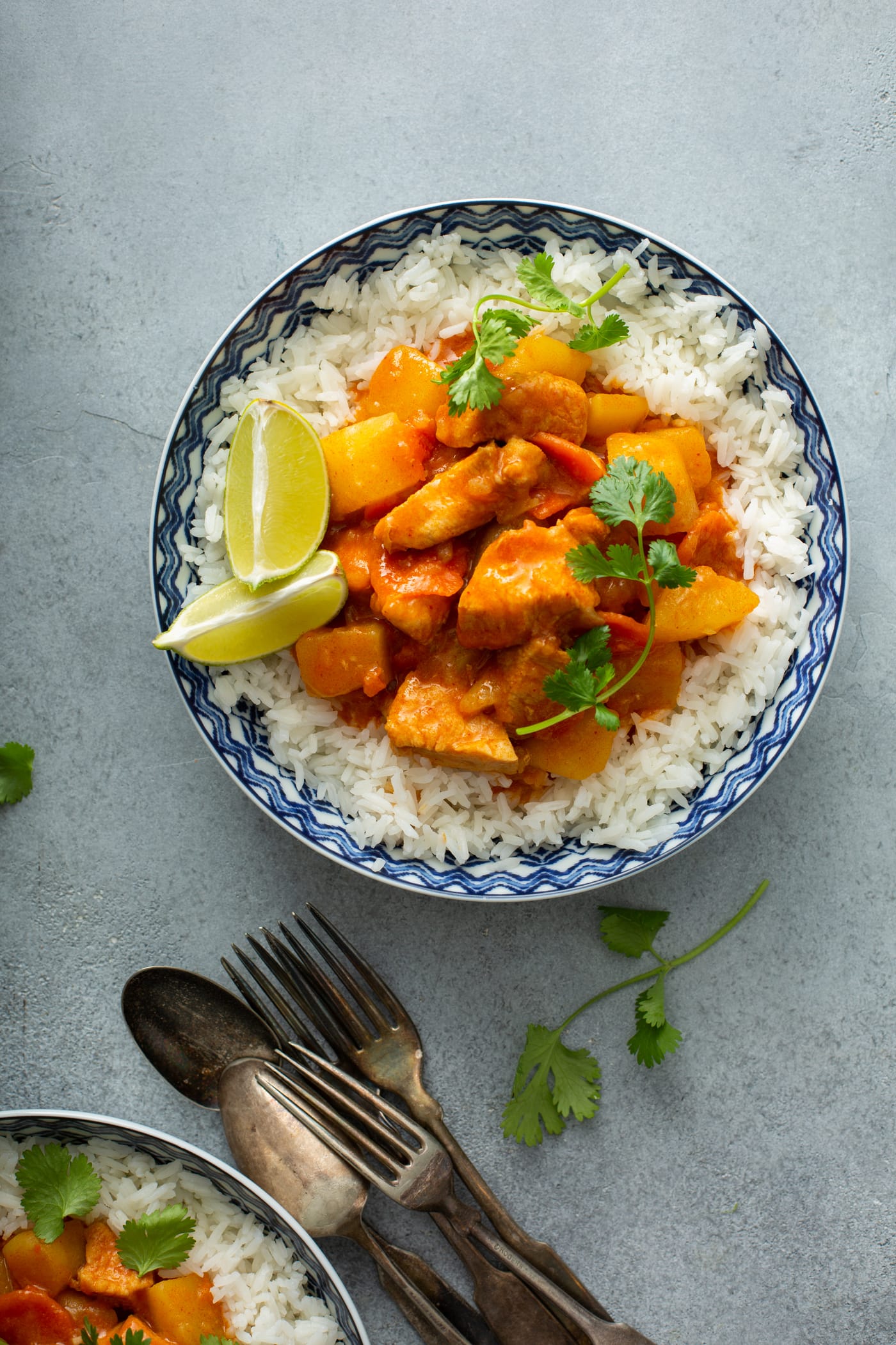 A bowl of rice with chicken curry on top. There are fresh parsley leaves sprinkled on top and two lime wedges on the edge of the rice. Spoons and forks are on the table next to the bowl.