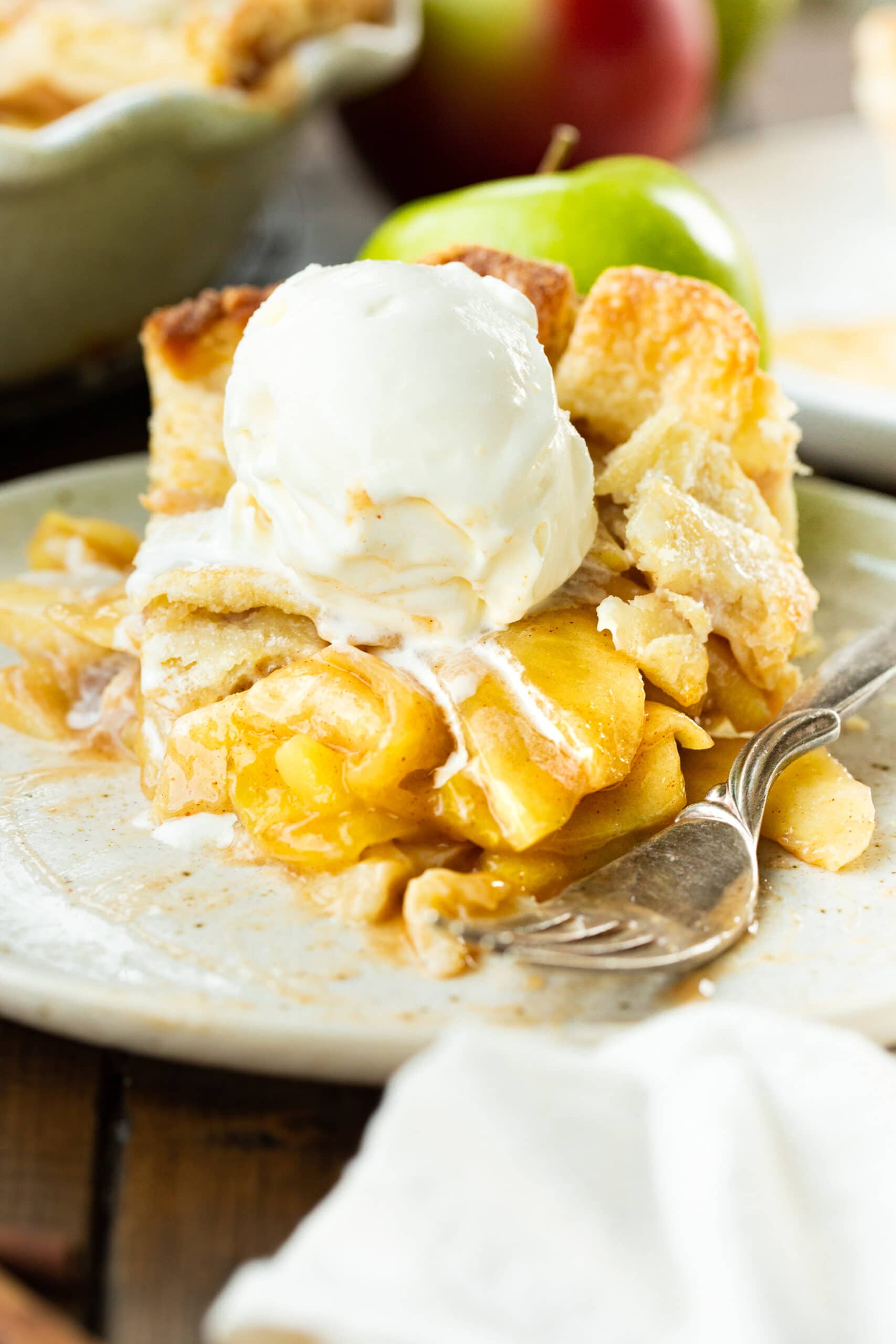 a photo of a slice of homemade apple pie sitting on a small dessert plate with a scoop of vanilla ice cream on top that is melting down the sides.