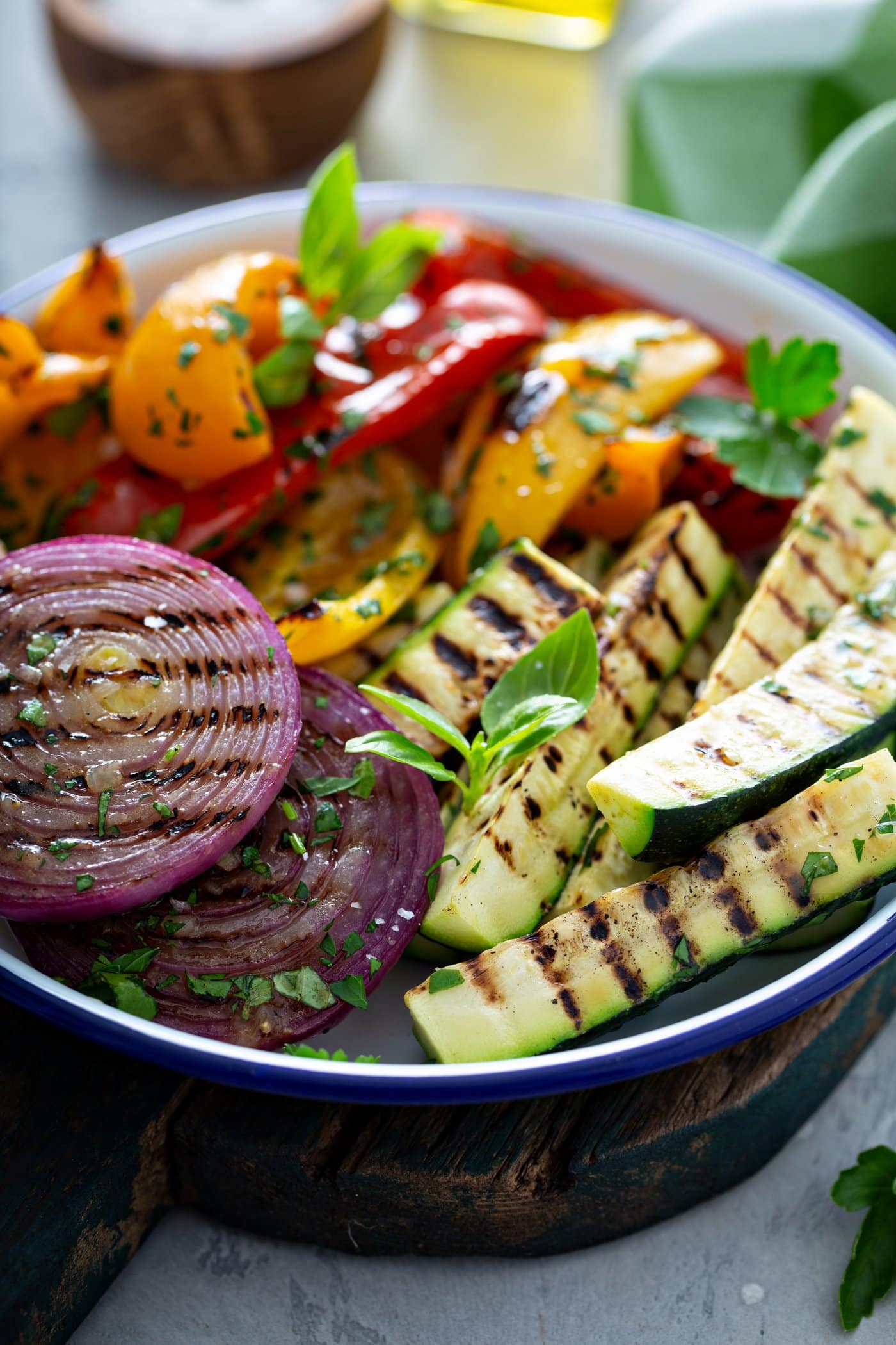 A serving dish of grilled veggies. The zucchini wedges, sliced red onions, and red and yellow bell peppers have perfect grill marks. and the veggies have a few parsley leaves scattered on top.