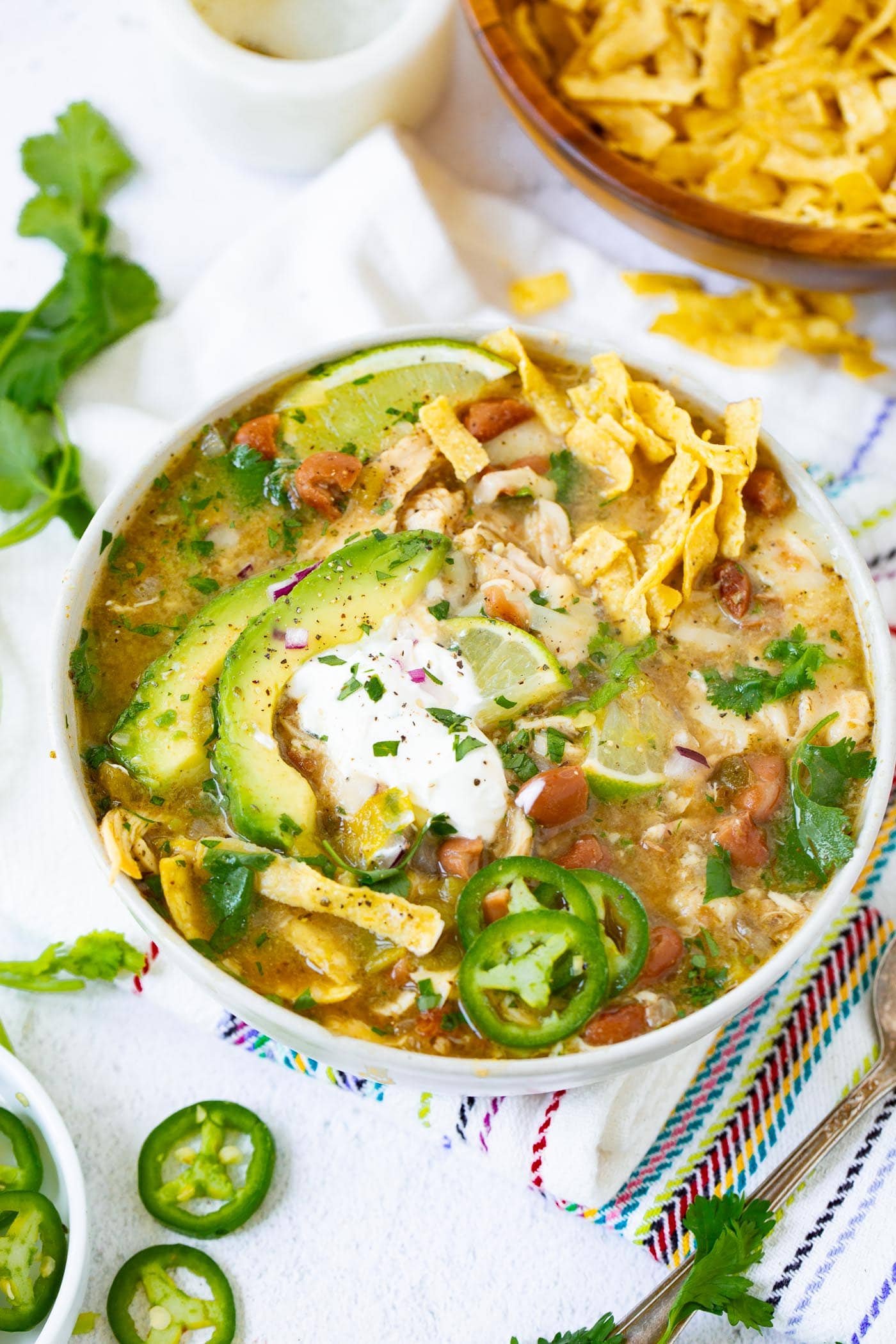 a photo of a bowl full of white chicken chili topped with a dollop of sour cream, avocado slices, tortilla chip strips and slices of fresh jalapeno.