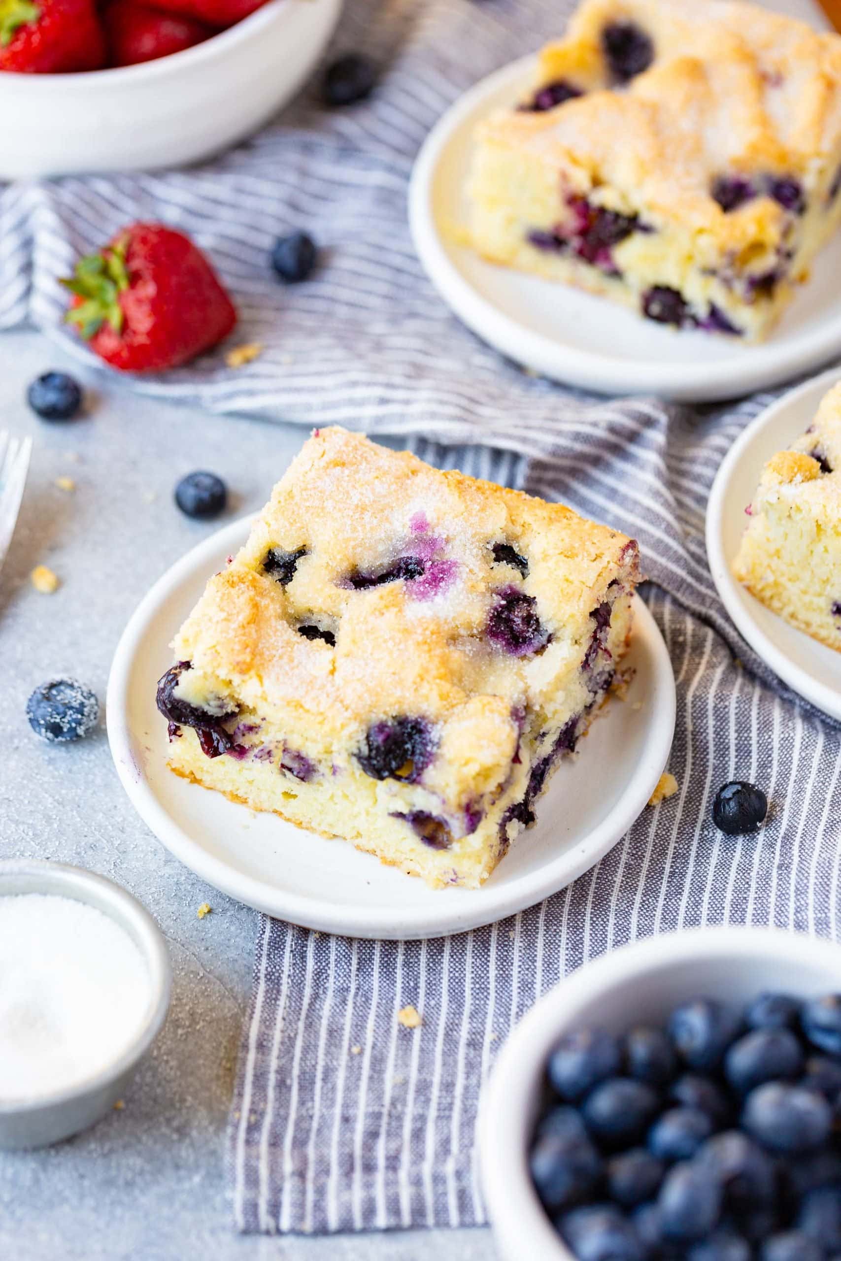 a photo of a piece of fluffy moist blueberry breakfast cake on a small white plate surrounded by other small plates full of breakfast cake.
