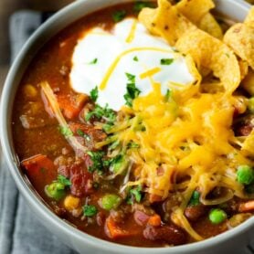 a photo of a bowl of taco stew topped with a dollop of sour cream, a pile of shredded cheddar cheese and a few Fritos chips.