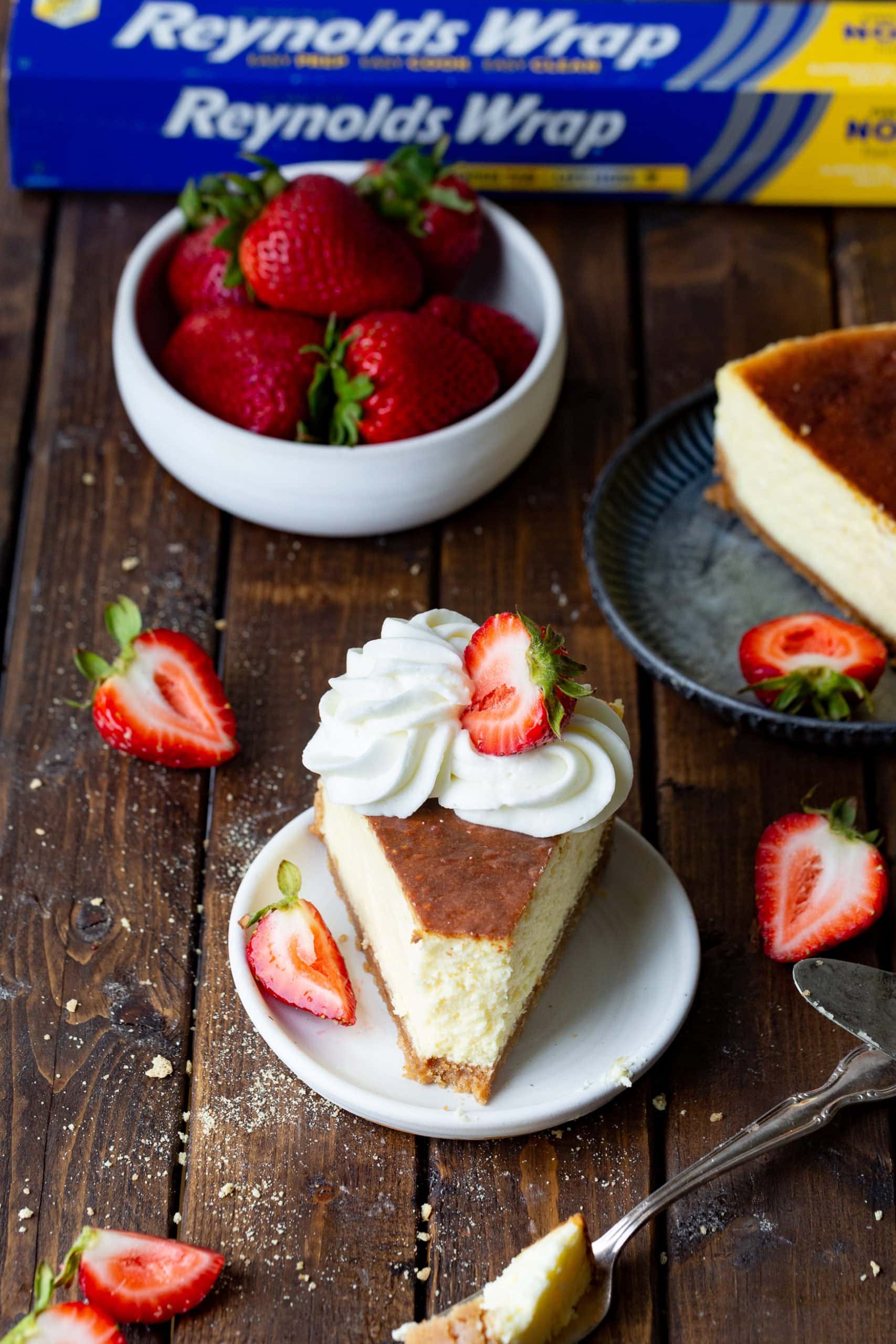 a photo of a slice of classic cheesecake topped with glazed whole strawberries and dollops of whipped cream on a small white dessert plate.