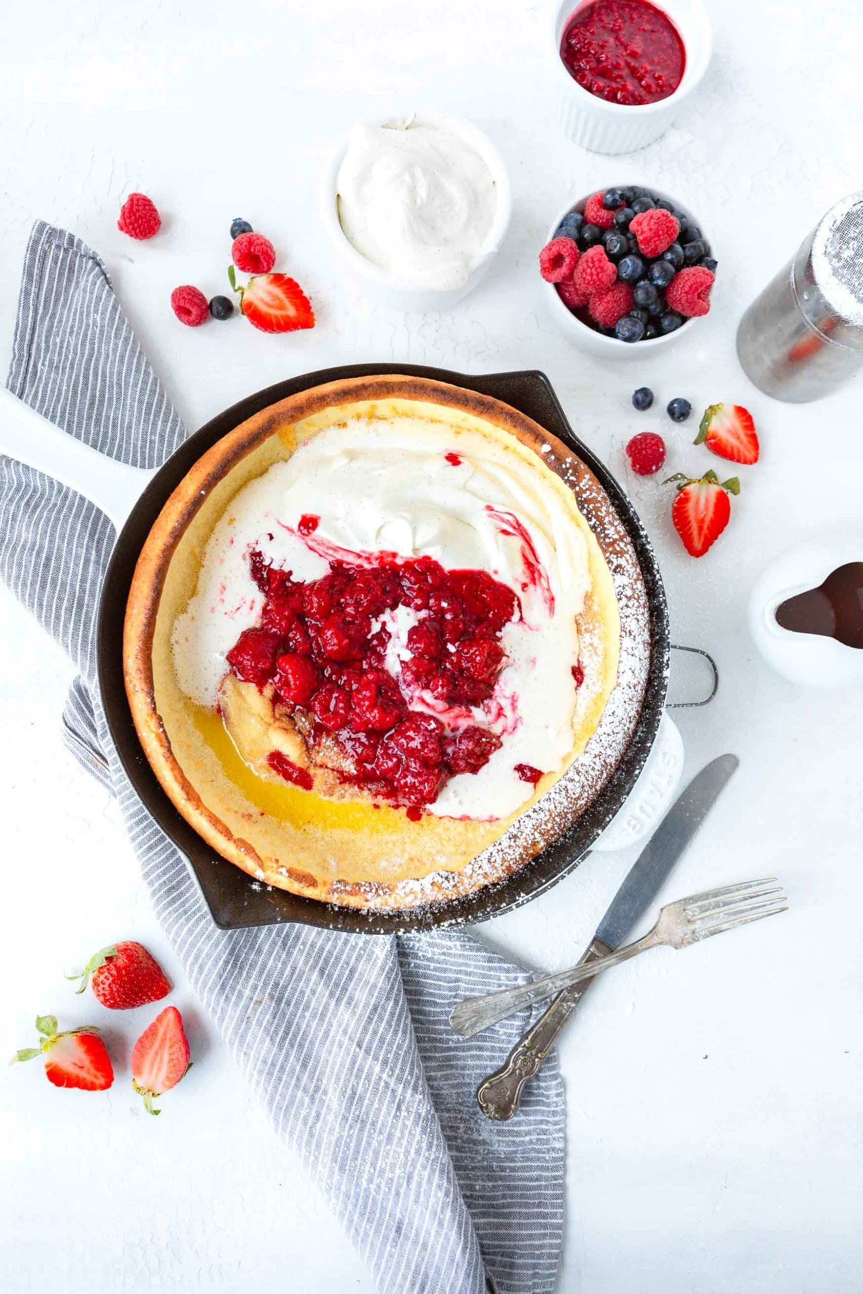 A photo of a baked dutch baby pancake topped with whipped cream and fresh berries with a cup of fresh berries sitting next to the pan.