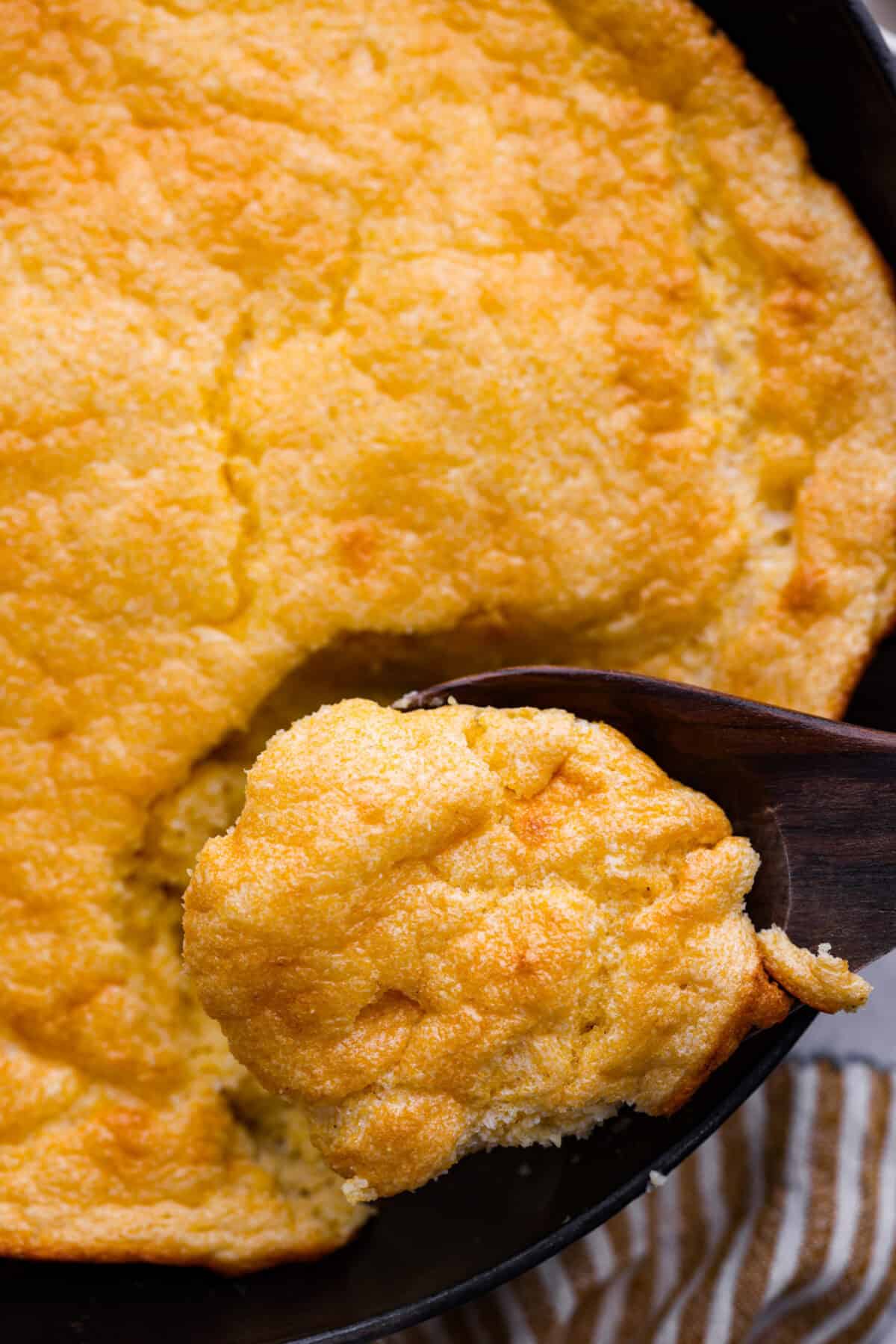 Close up shot of a scoop of spoon bread in a wooden spoon over the skillet. 