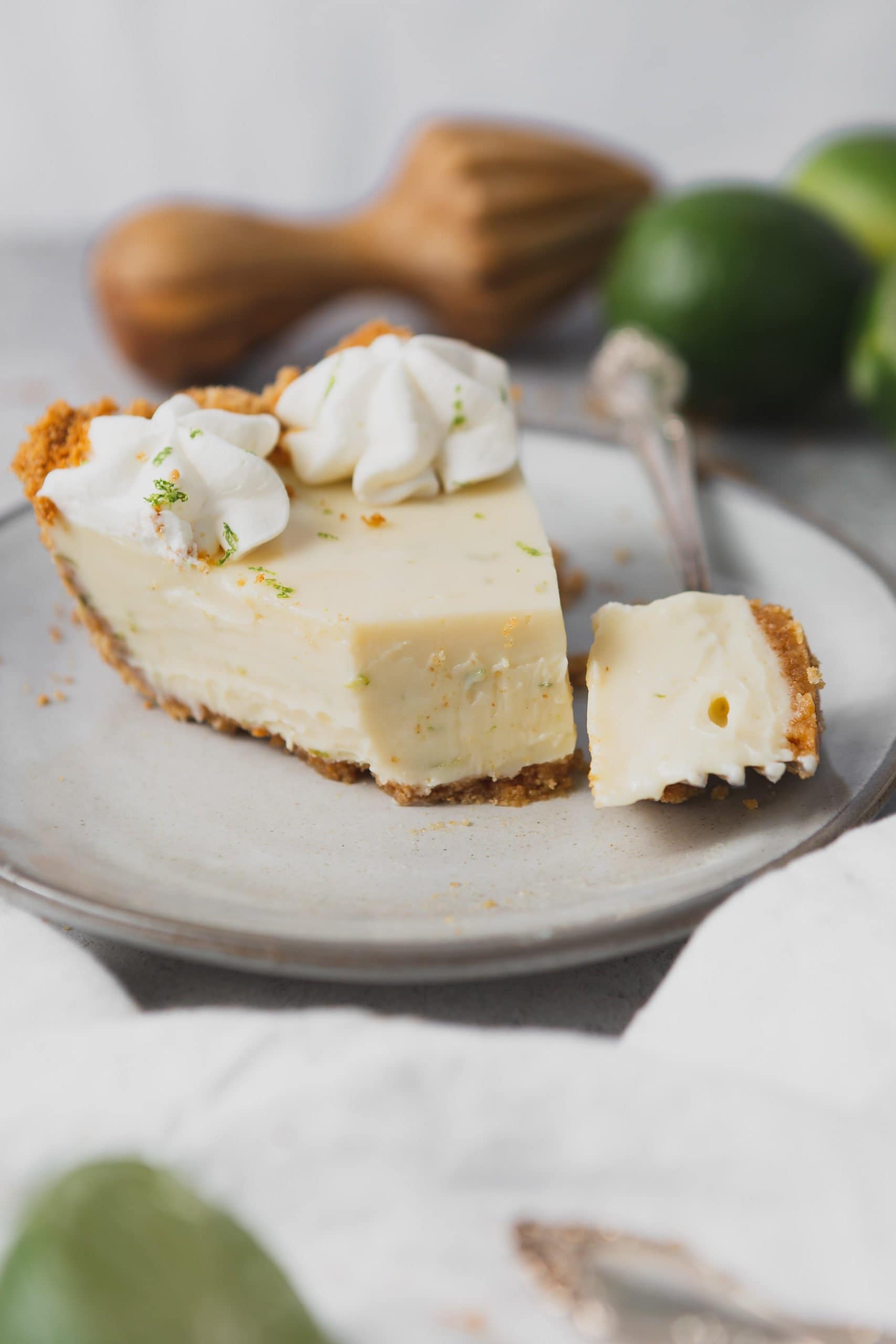 A slice of key lime pie with a piece cut from it with a fork. It is on a white dessert plate and has two dollops of whipped cream and a little lime zest on top.