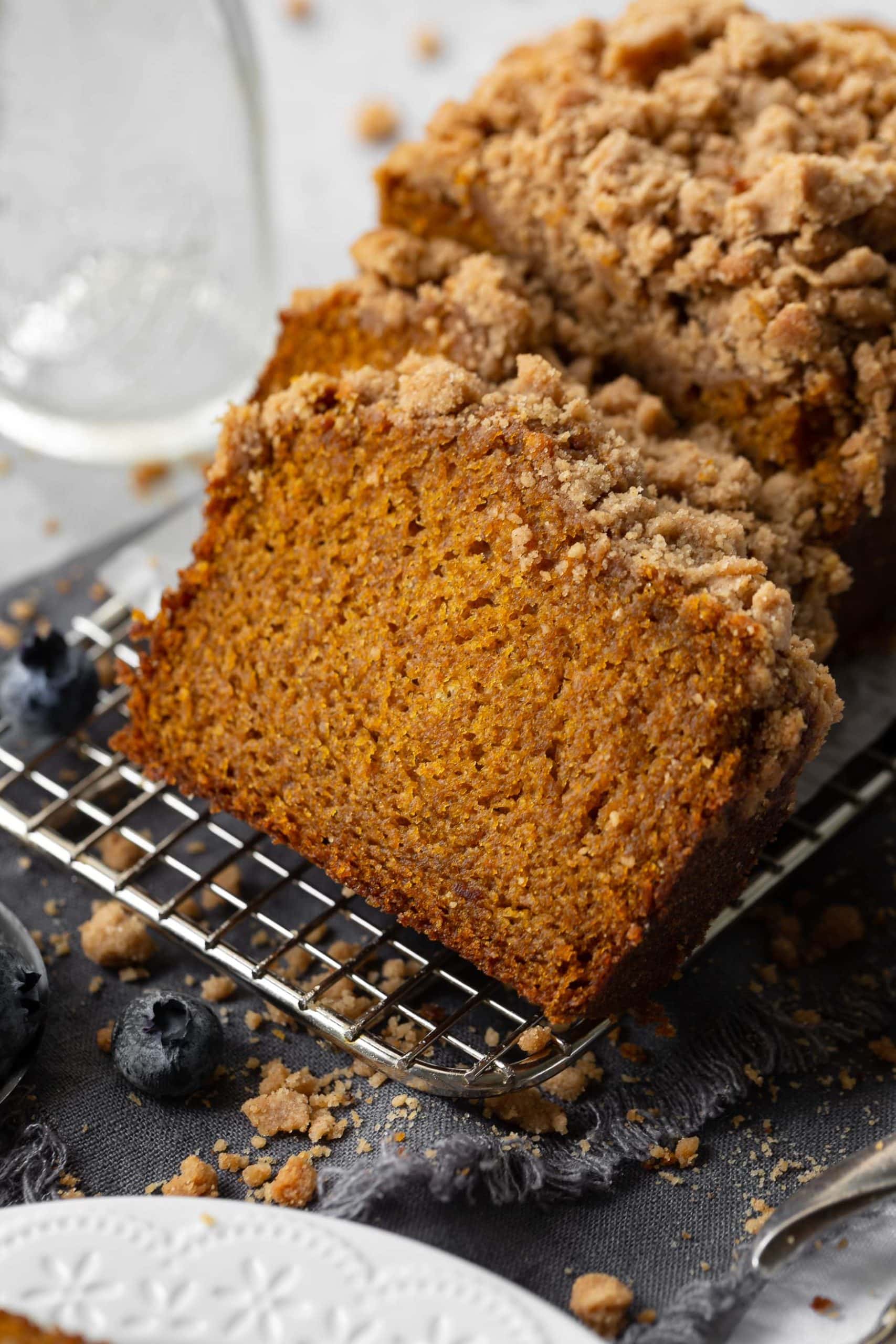 a photo of several slices of pumpkin bread topped with cinnamon streusel leaning up against half a loaf of uncut bread all sitting on a wire cooling rack.