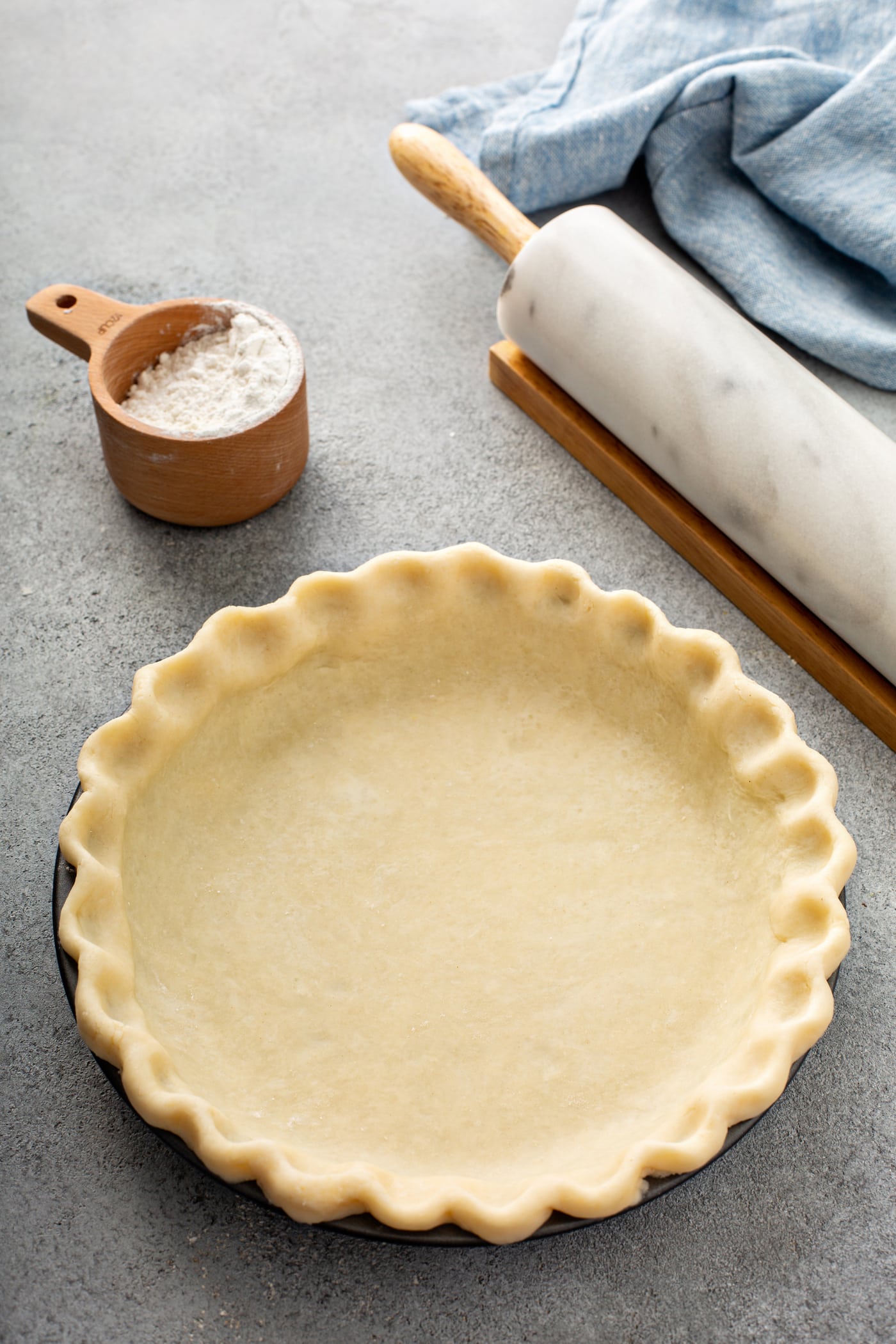 A photo of an uncooked butter pie crust in a pie tin sitting on a table next to a rolling pin and a cup of flour.
