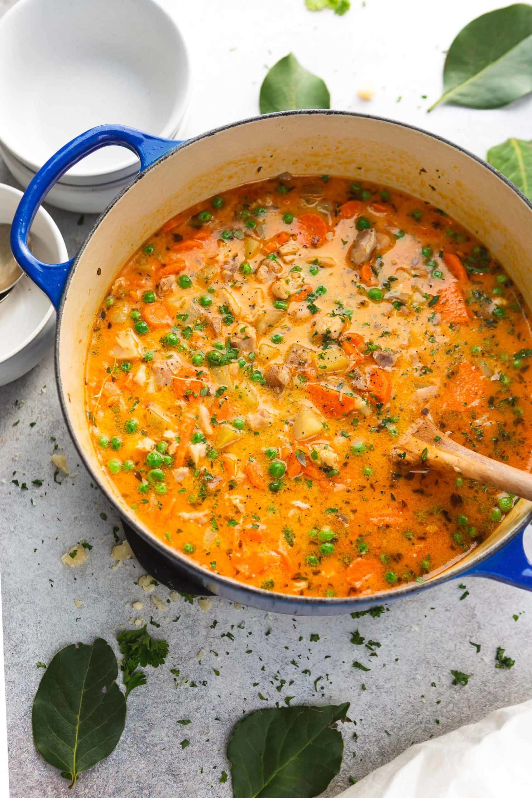 A pot of chicken stew. The stew is thick and bright colored with bits of celery, herbs, chicken and potatoes. A wooden spoon is in the pot and chopped herbs are on the counter top next to the pot.