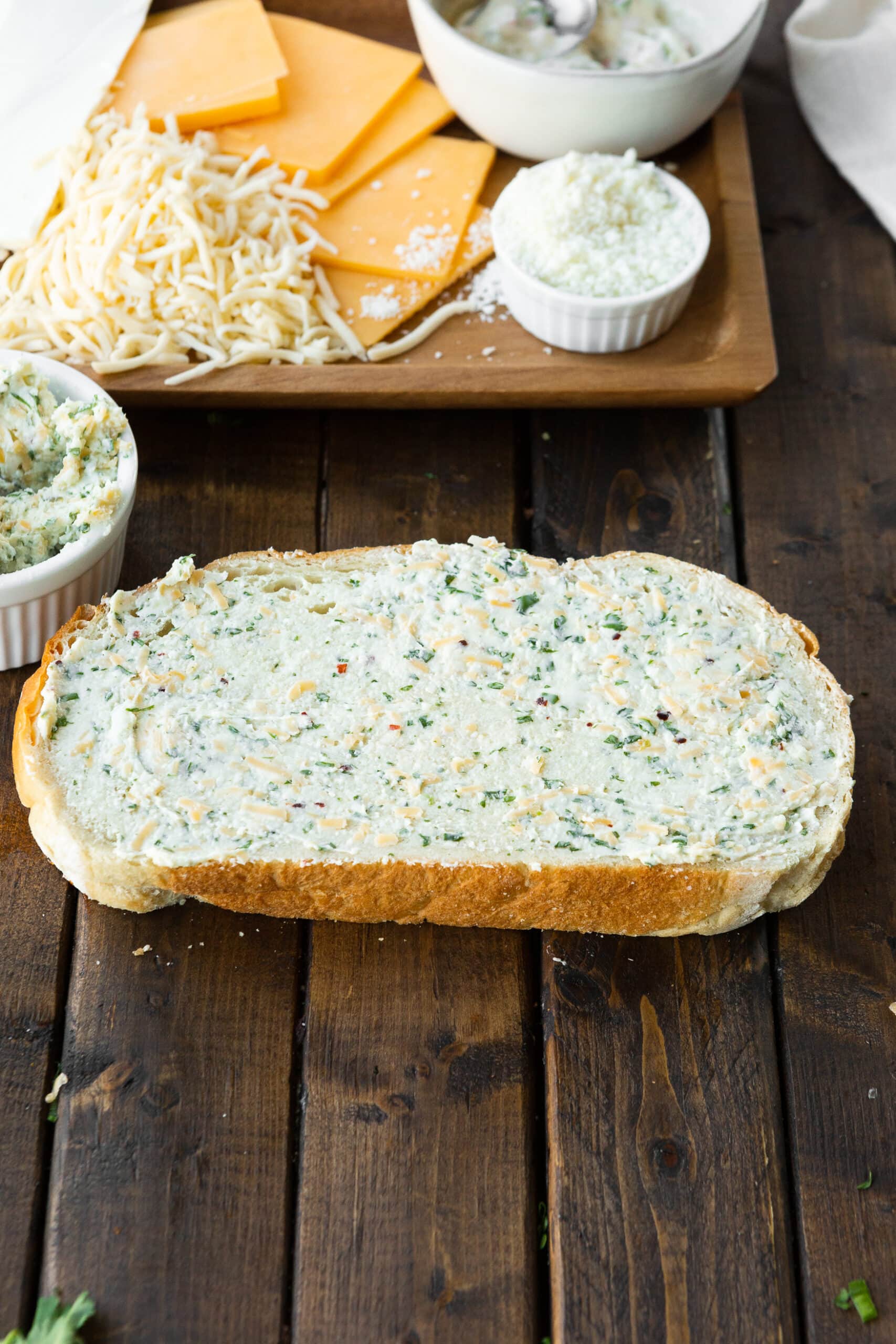a photo of a single slice of sourdough bread that has been covered with a flavored butter spread with a platter of cheese in the background.