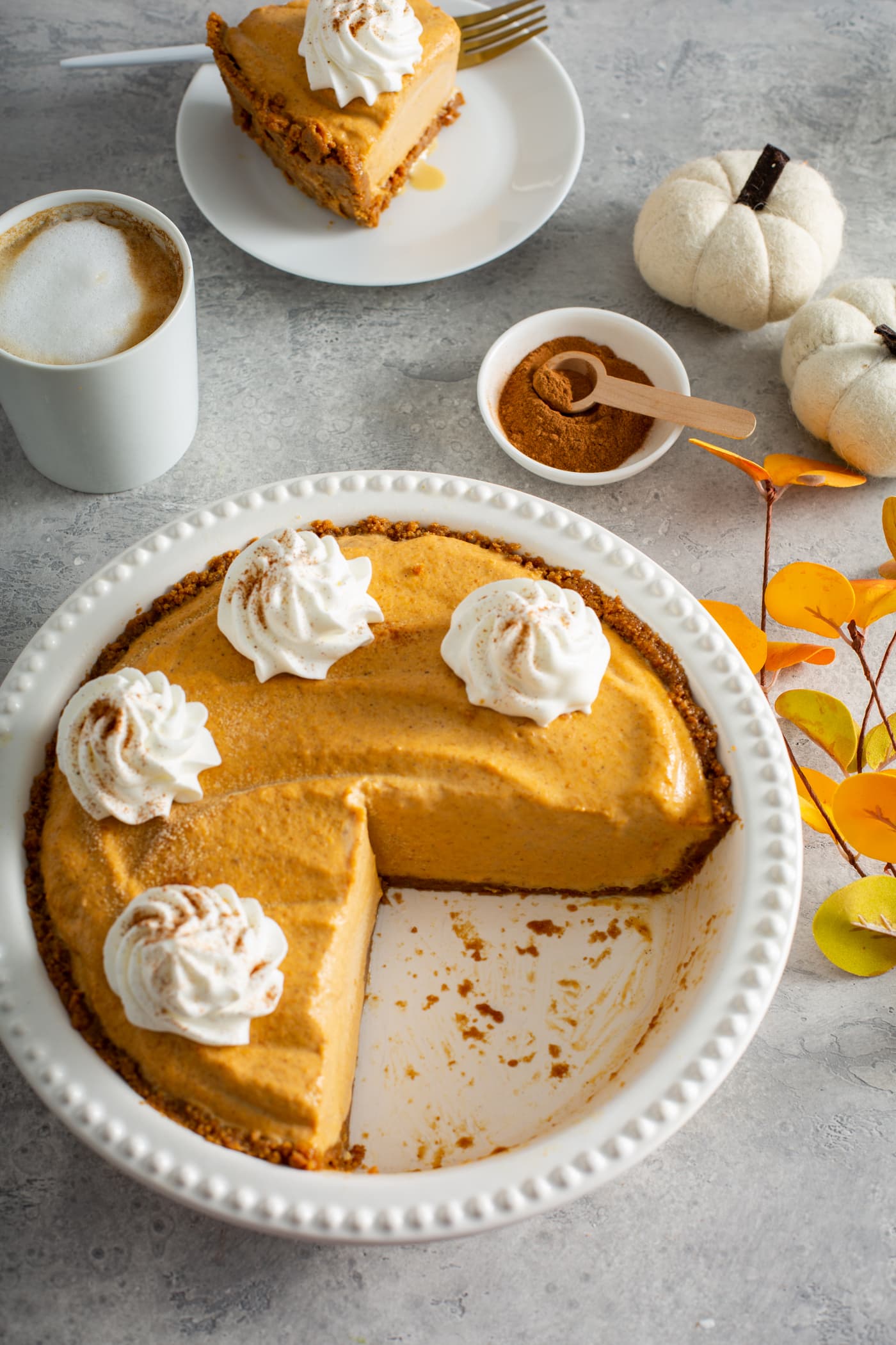 A white pie dish with part of a pumpkin ice cream pie that is topped with dollops of whipped cream sprinkled with cinnamon. There Is A white mug with a drink topped with whipped cream, a small dish with a wooden spoon filled with cinnamon, two tiny white pumpkins and a dessert plate with a slice of pie in the background.