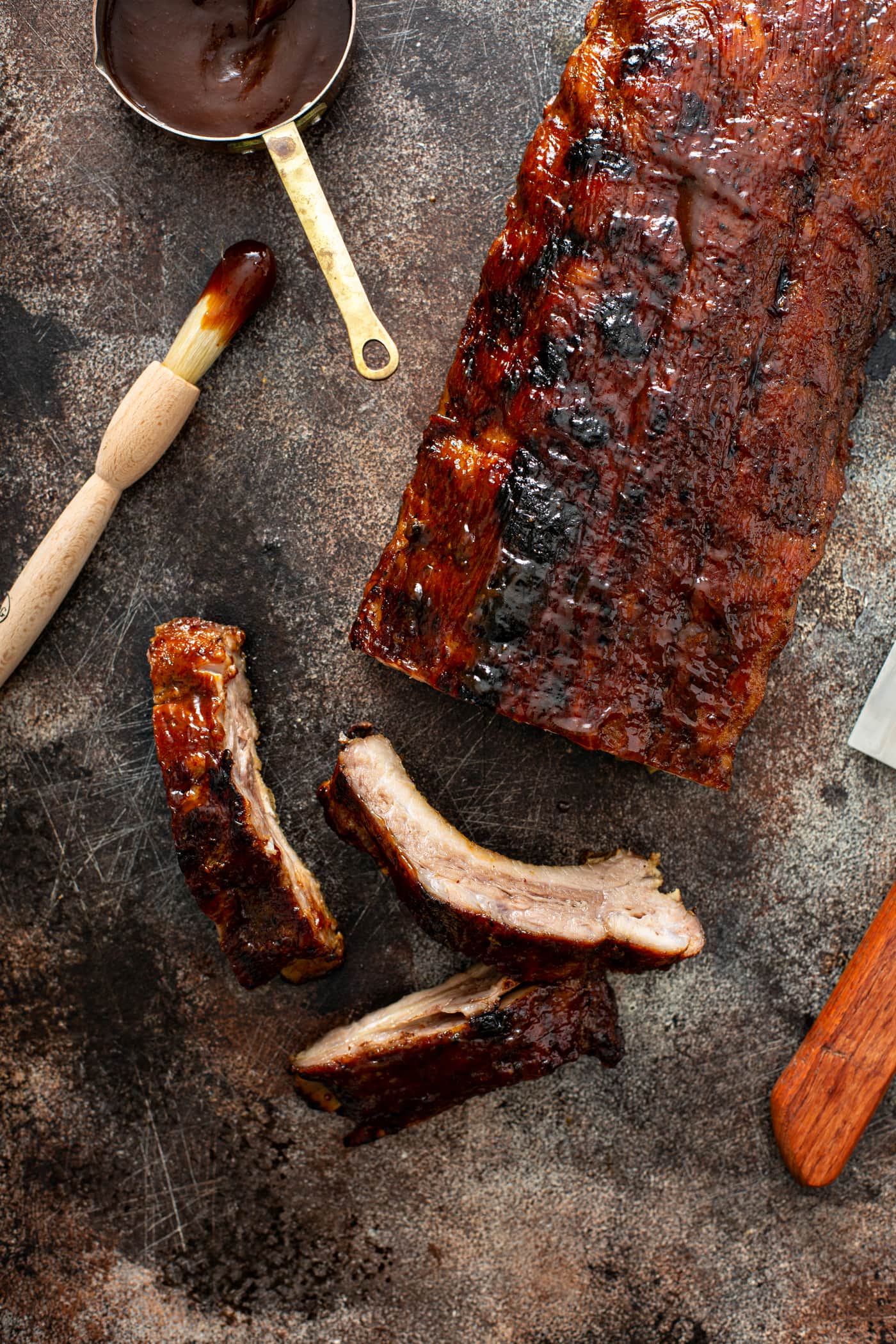 A rack of grilled ribs with three ribs sliced from the rack. A brush with glaze for the ribs is next to the rack of ribs.