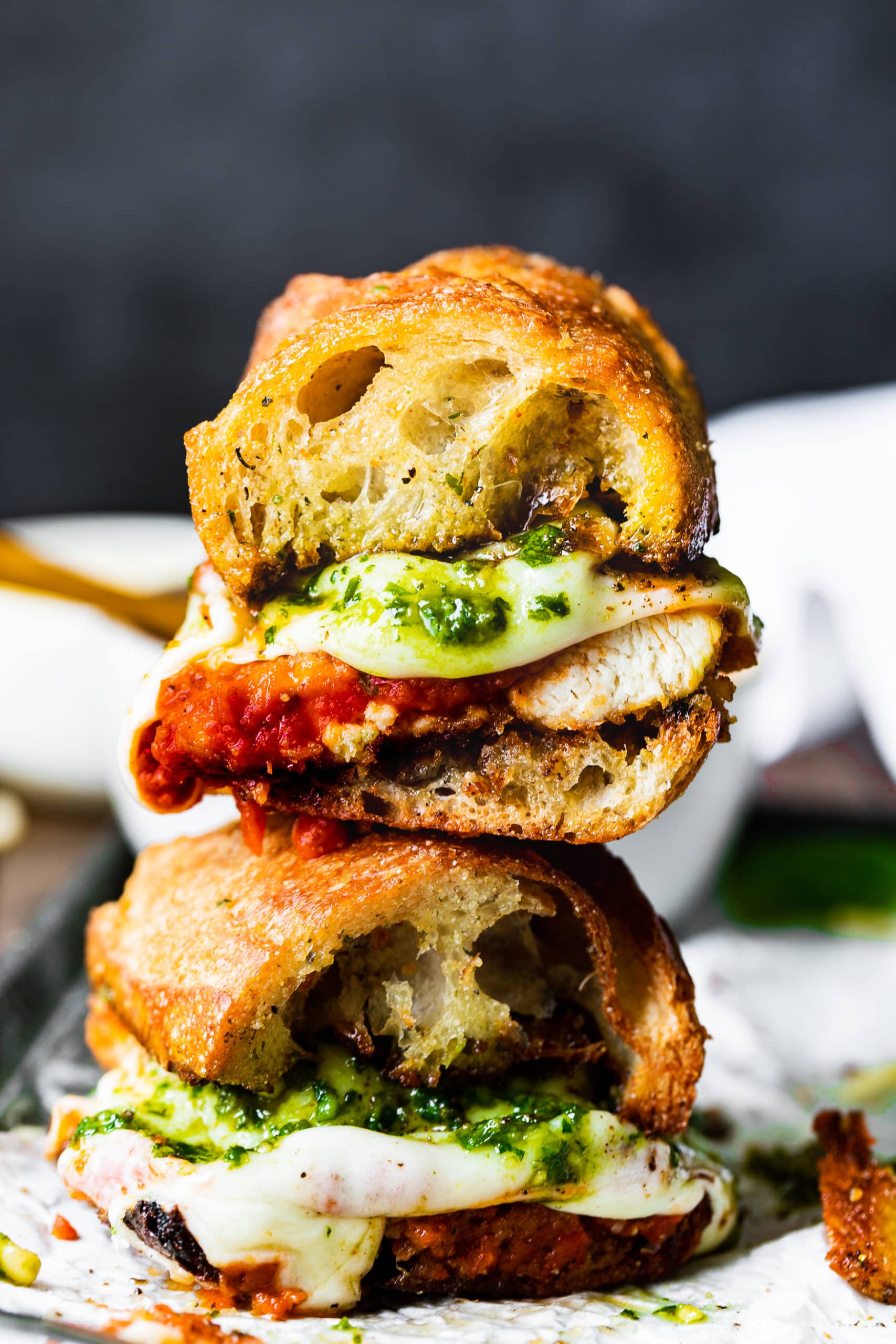 a photo of two small chicken parm sandwiches stacked on top of each other complete with juicy chicken breasts, marinara, melty mozzarella and pesto.