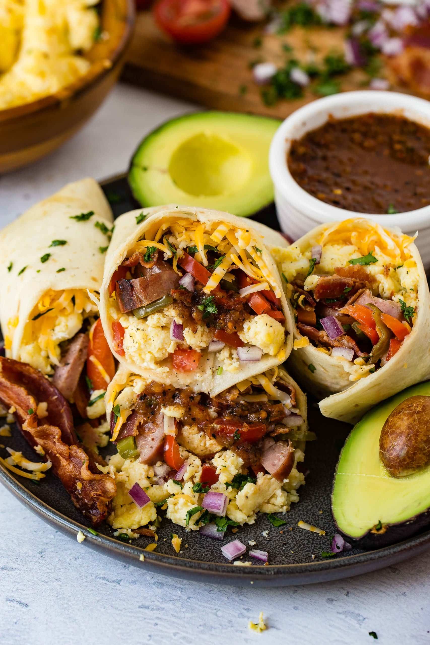A plate of breakfast burritos. The burritos are stuffed with scrambled eggs, bacon, sausage, and peppers. There is a bowl of salsa on the side, and an avocado that is cut in half.