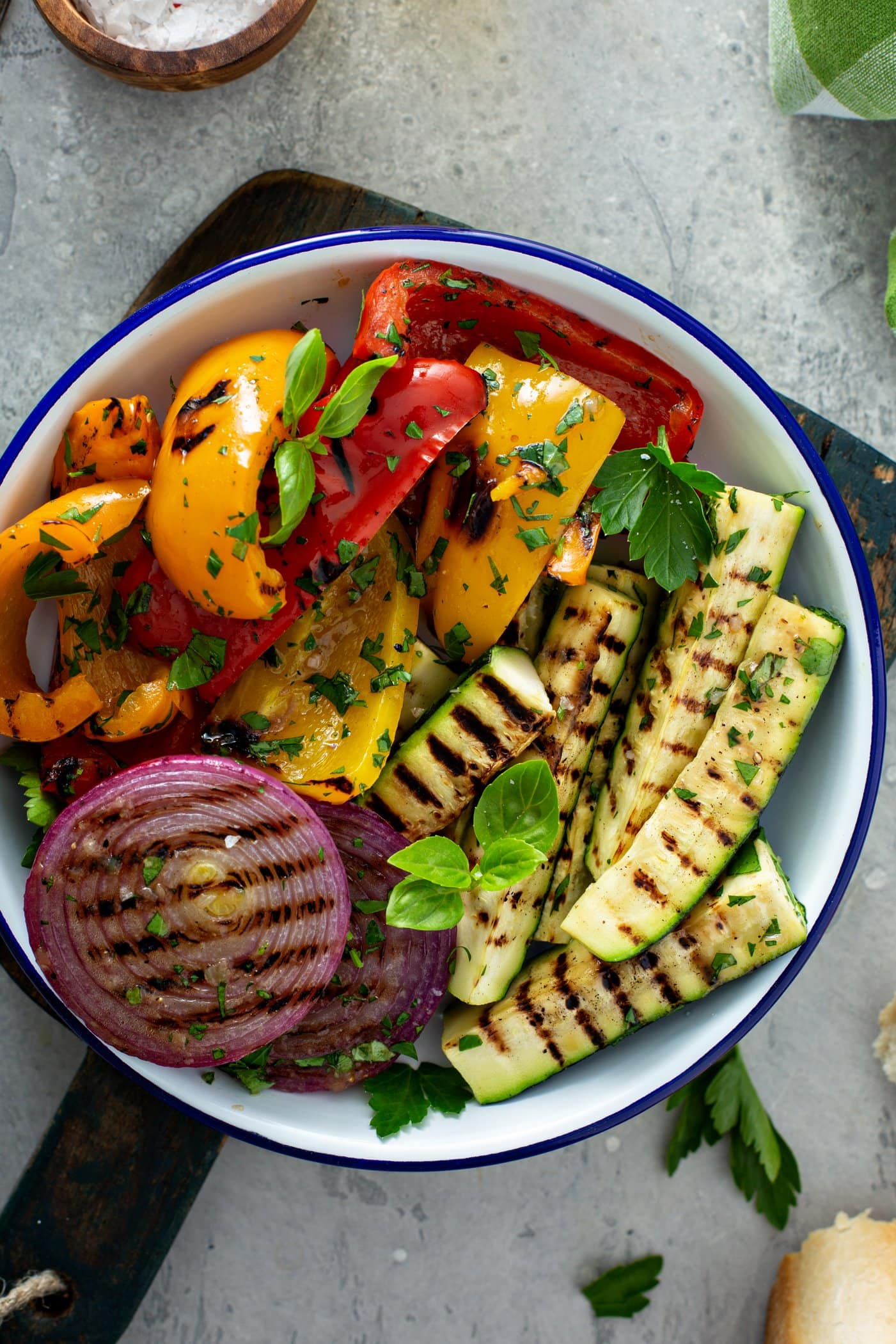 A serving dish of grilled veggies. The red onion slices, zucchini wedges and sliced red and yellow bell peppers have light brown grill marks and the veggies are sprinkled with parsley leaves.