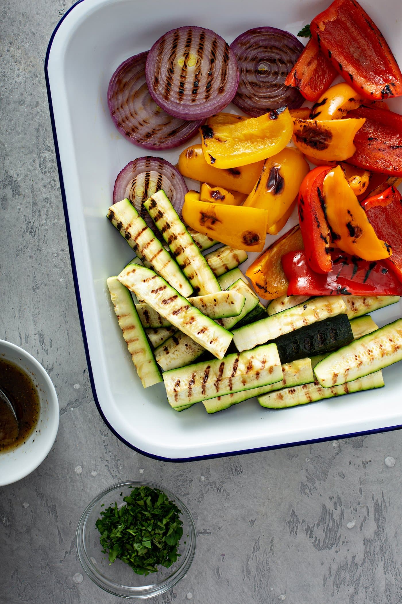 A plate of grilled veggies with a cup of balsamic glaze sitting next to the plate. The grilled zucchini wedges, slices of red onion and red and yellow bell peppers have lovely grill marks on them.