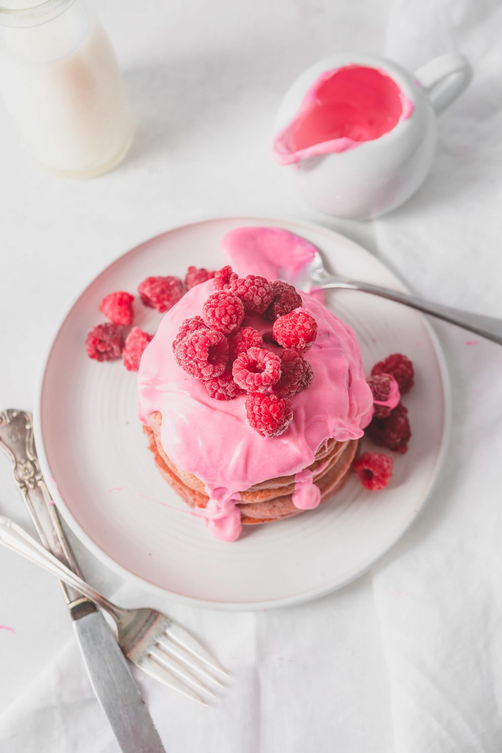 An overhead view of a stack of pink pitaya pancakes on a white plate. The pancakes are covered with a thick pink chocolate sauce topped with fresh raspberries. There is a spoon full of pink sauce on the plate and a container full of sauce in the background.