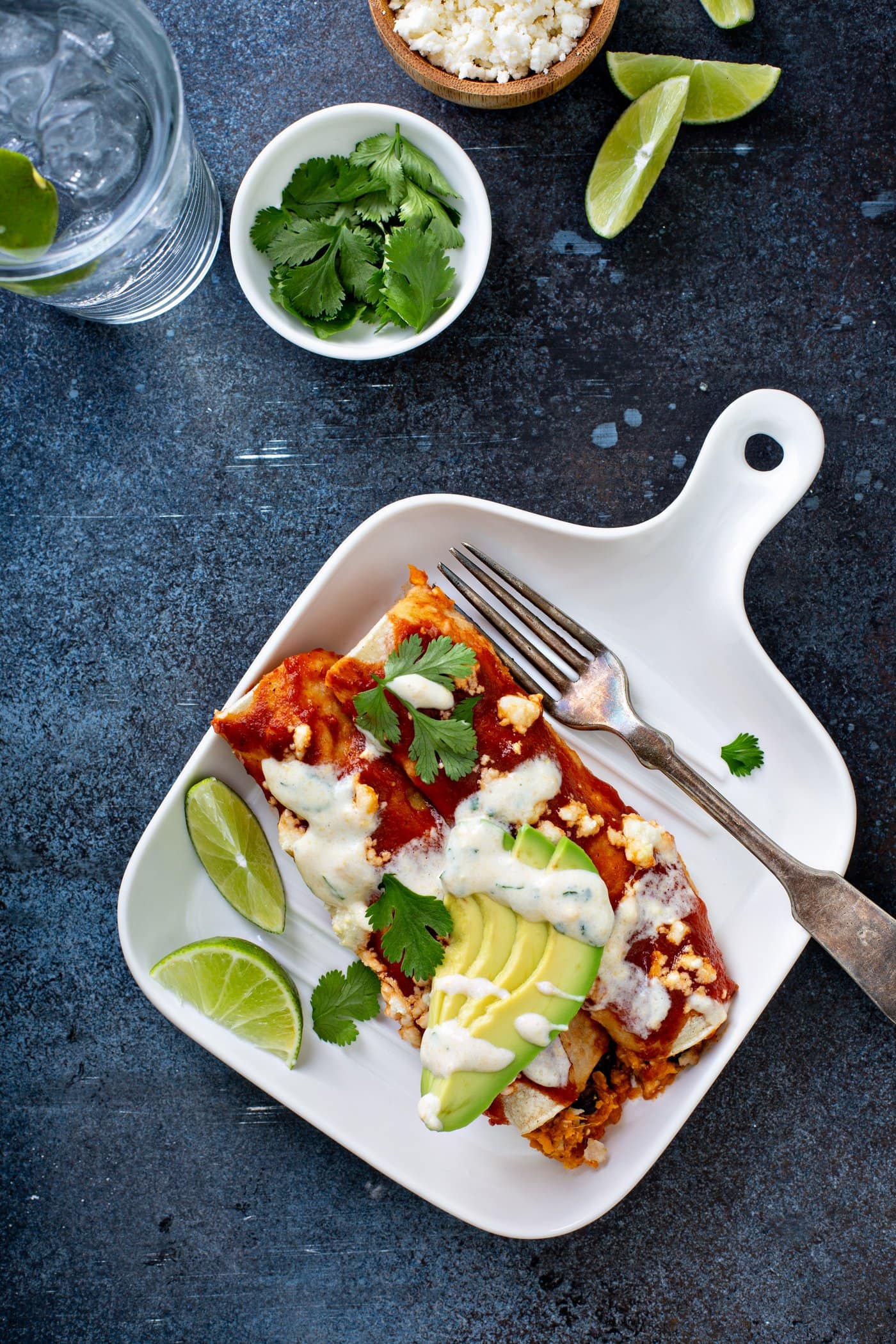 A dinner plate with sweet potato enchiladas and a fork. the enchiladas are topped with red enchilada sauce, sliced avocado, cilantro leaves, and a drizzle of sweet corn crema. There are two lime wedges next to the enchilada. A cup of cilantro and a glass of water are in the background.