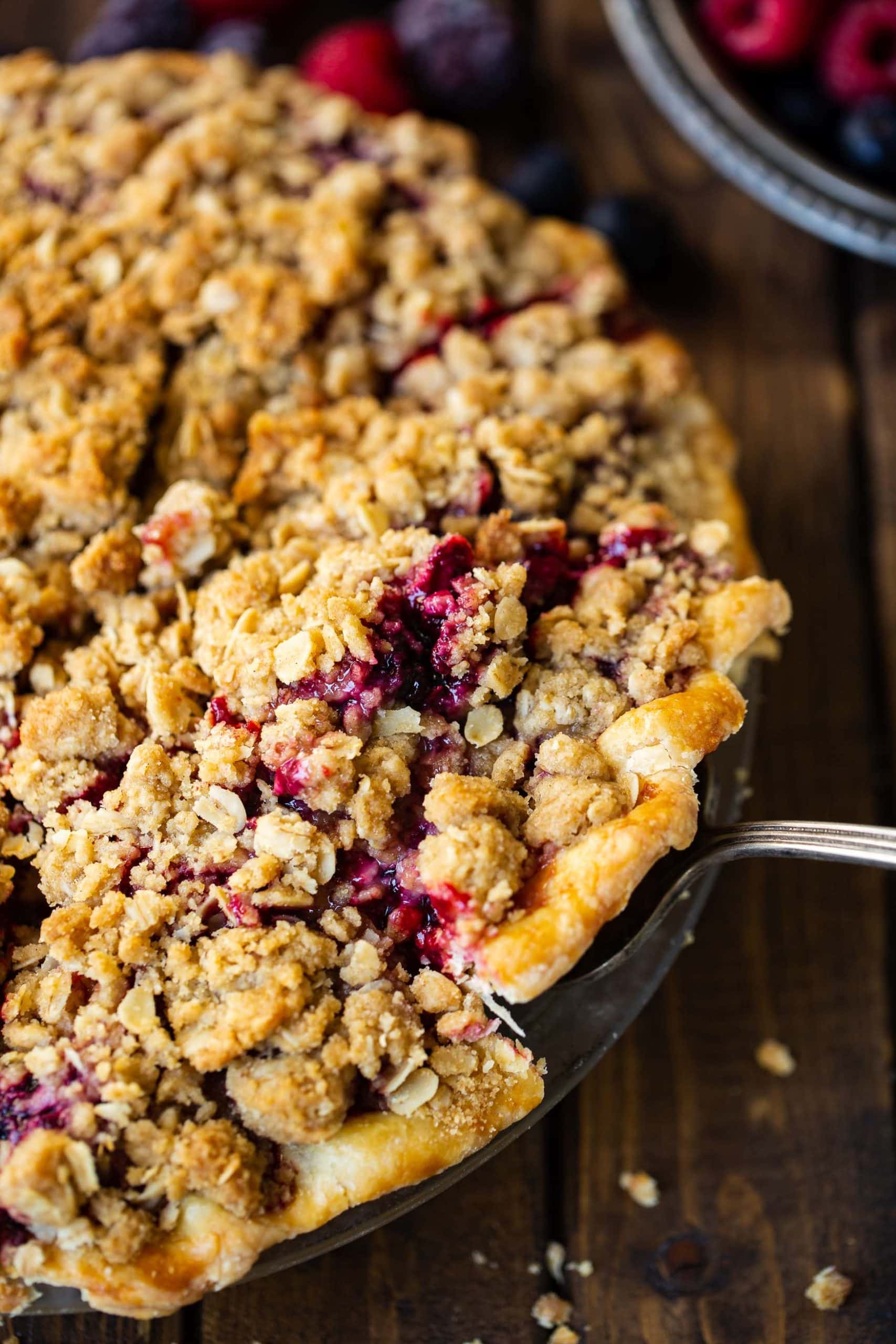a photo of a single slice of mixed berry pie with streusel topping being removed from a whole pie.