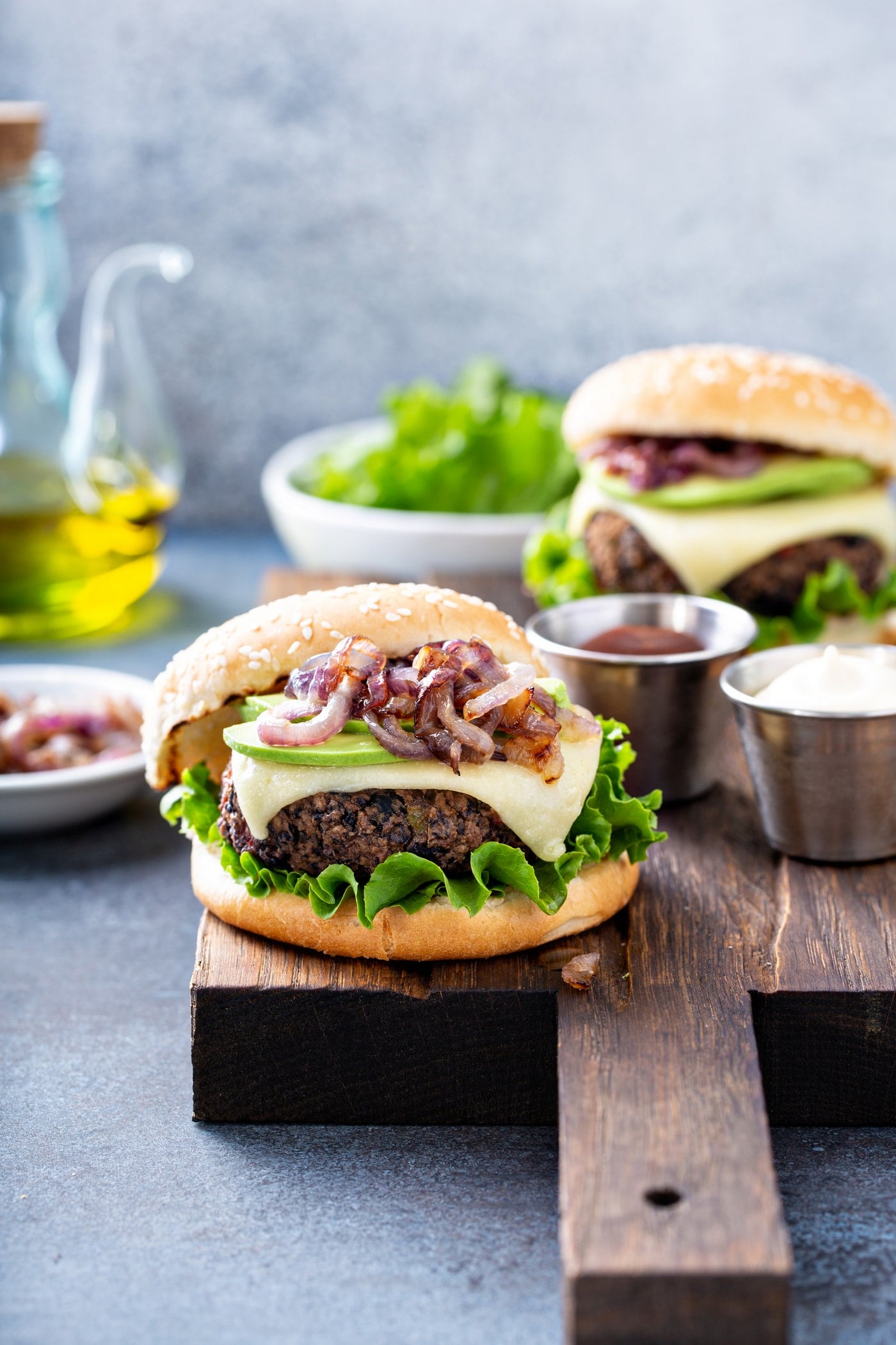 A black bean burger on a sesame seed bun topped with onions, avocado, cheese, and lettuce. There is another burger in the background and a bowl of lettuce, a container of oil, a dish of sliced onions, and a container of mayo.