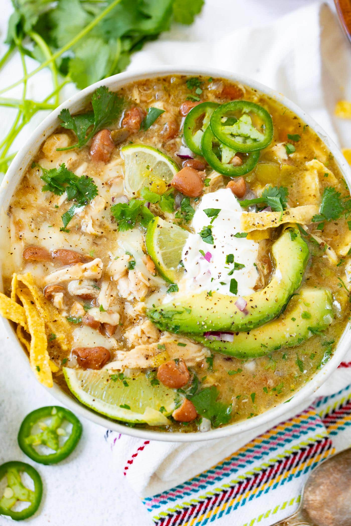 a photo of a bowl full of white chicken chili topped with a dollop of sour cream, avocado slices, tortilla chip strips and slices of fresh jalapeno.