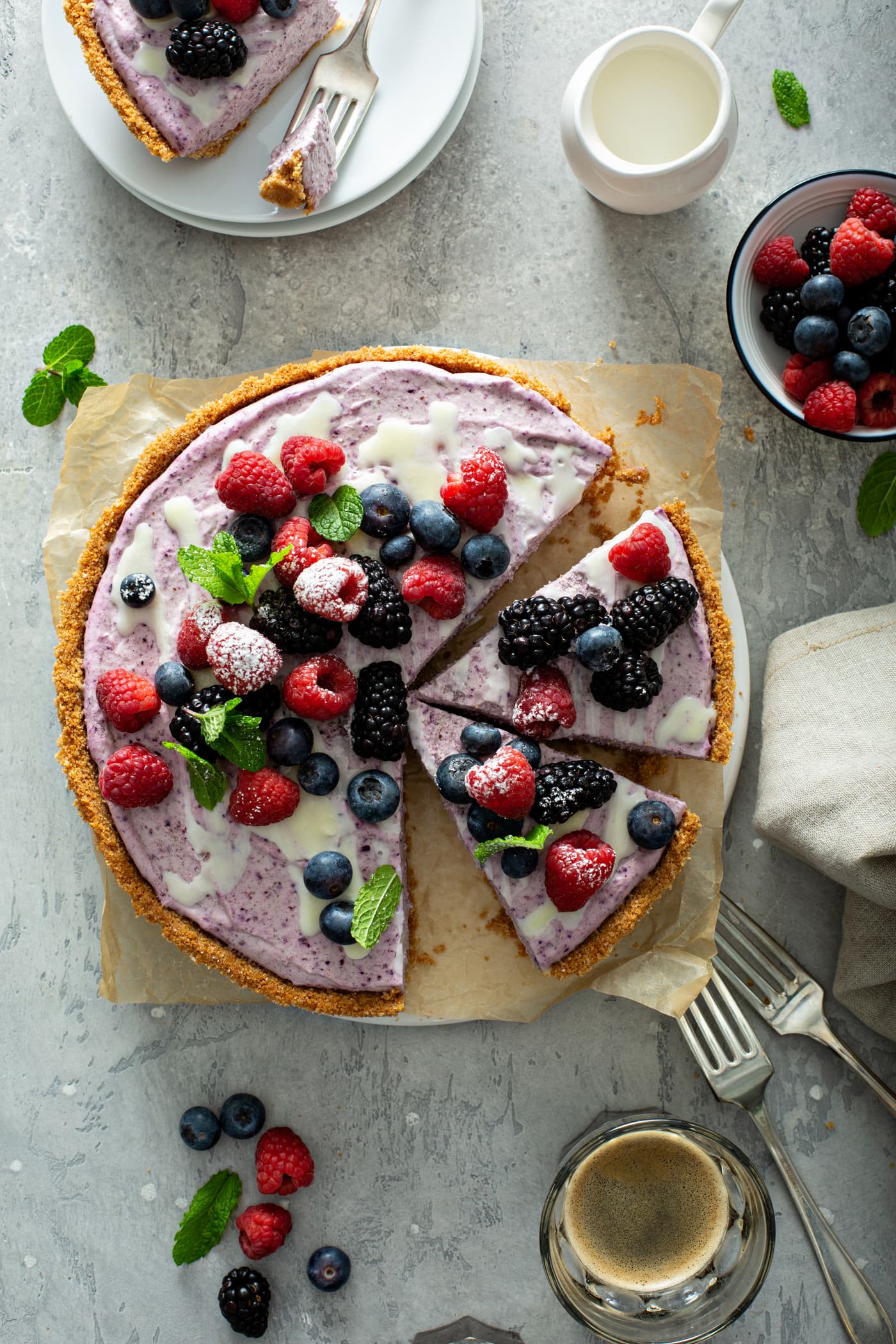 A berry white chocolate pie that has been sliced. A slice has been removed and two more slices are separated. The pie has a graham cracker crust and is topped with strawberries, blue berries and raspberries. A few mint leaves are scattered on top of the pie and on the table next to the pie. There are two forks, a napkin and a bowl of mixed berries on the table.