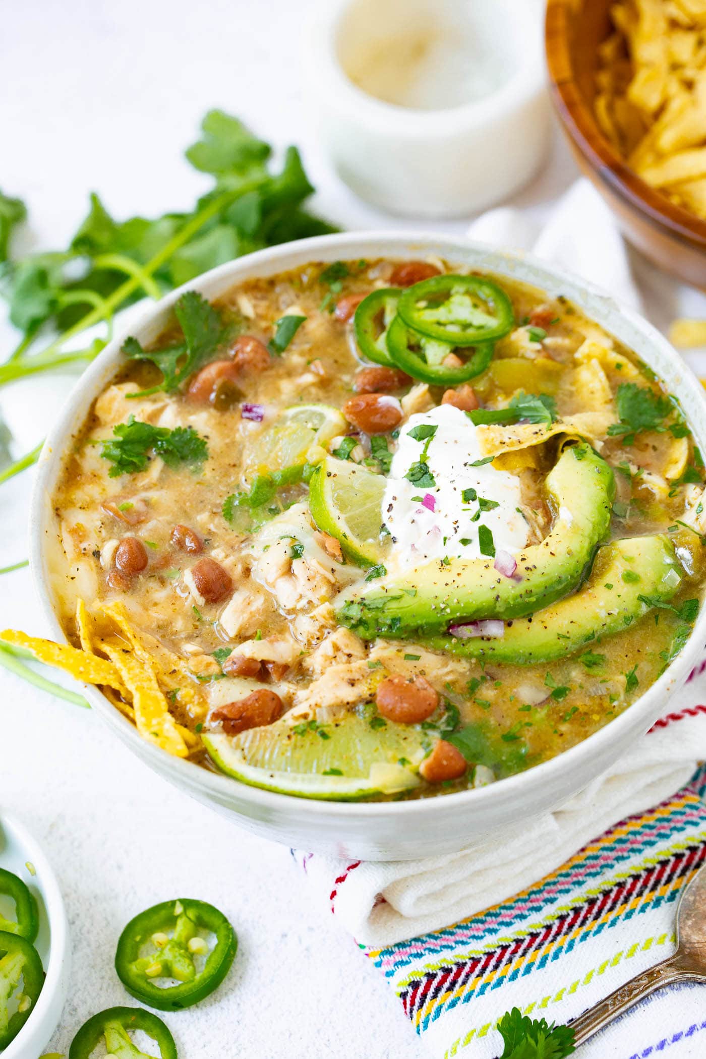 a photo of a bowl full of white chicken chili topped with a dollop of sour cream, avocado slices, tortilla chip strips and slices of fresh jalapeno.