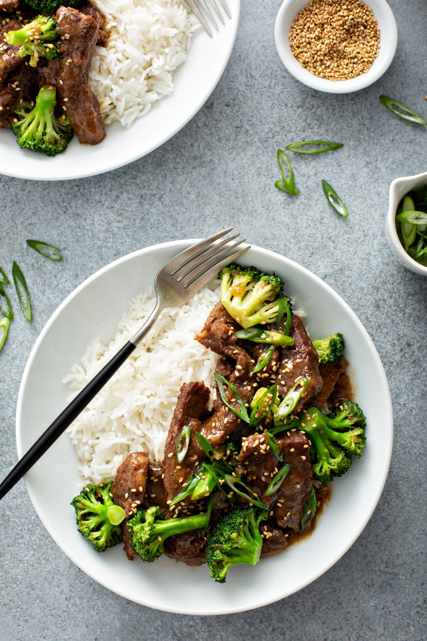 A photo of a white bowl full of beef and broccoli and a side of white rice with a fork sitting on top of ther rice.