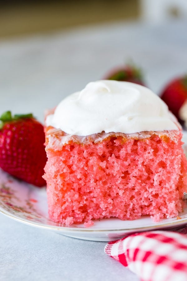 A floral china plate with a slice of pink strawberries and cream jello cake with a little cool whip on top and strawberries behind it.