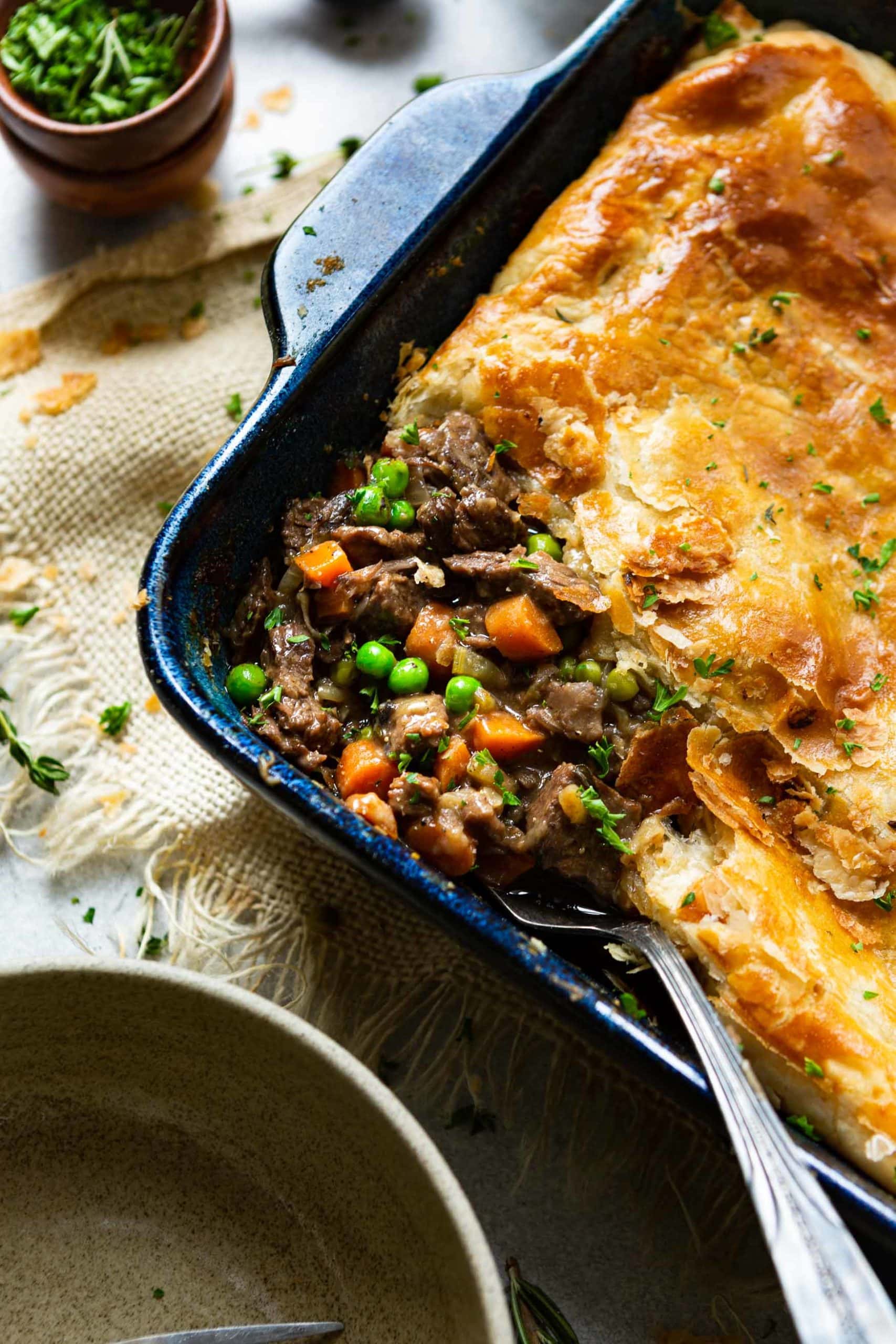 A photo of a blue ceramic rectangular baking dish full of a beef pot pie topped with golden puff pastry. a serving of the pot pie has been removed and the filling has filled in the missing piece and the serving spoon is sitting in the dish.