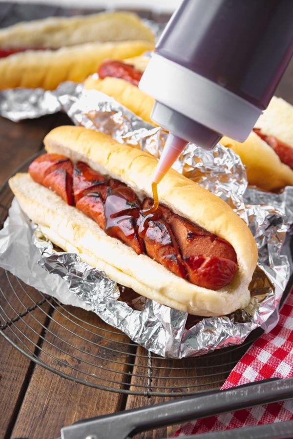 a photo of a grilled hotdog sitting in a bun inside of aluminum foil being drizzled with a sauce from a plastic bottle.