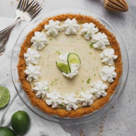 A key lime pie in a graham cracker crust. Dollops of whipped cream are around the edge of the pie and two slices of lime are in the middle. Limes and forks are beside the pie.