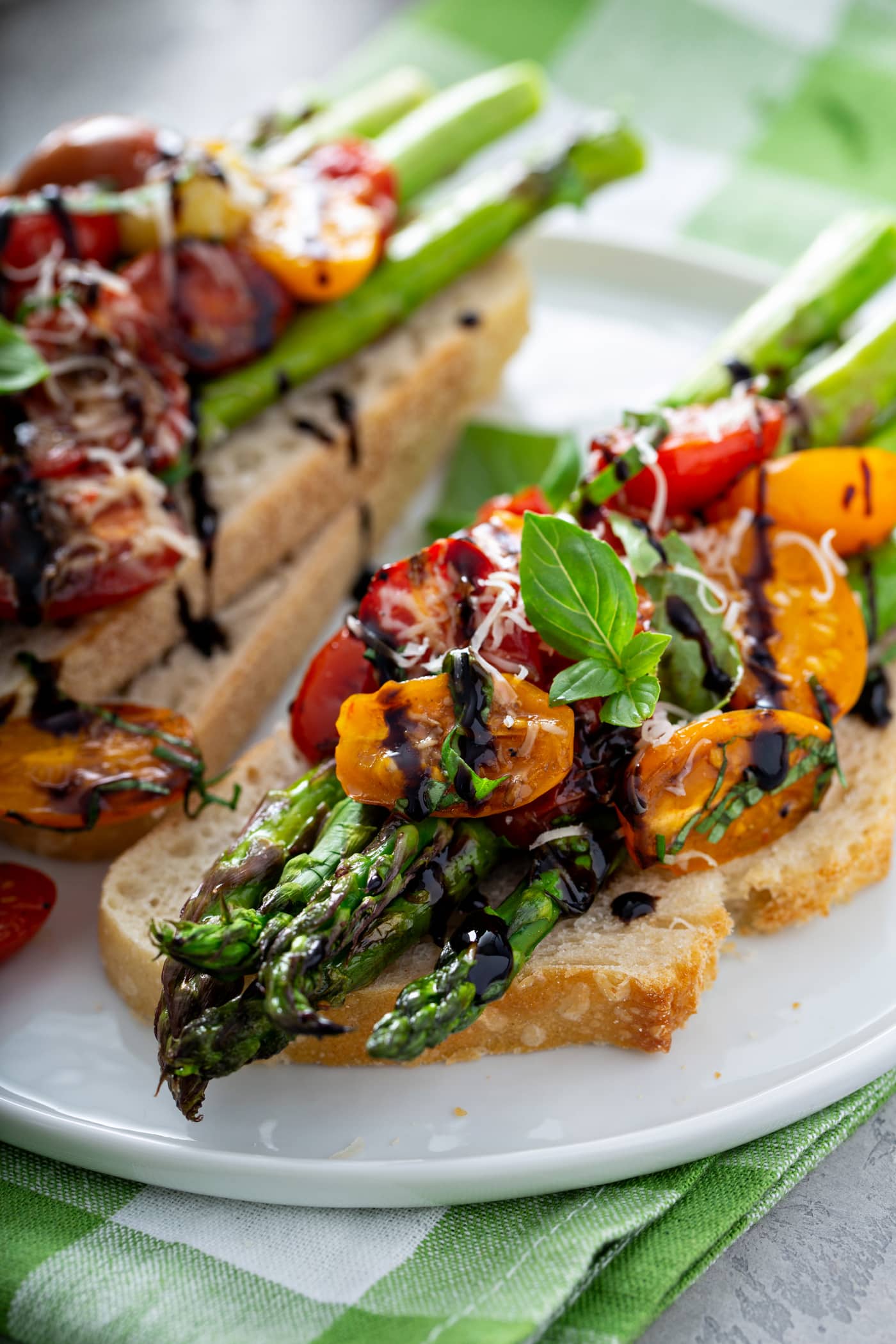 A serving of balsamic glazed roasted tomatoes and asparagus on a slice of bread. the roasted tomatoes and asparagus are drizzled with balsamic glaze and sprinkled with grated Parmesan cheese. There is another serving on two slices of bread in the background.