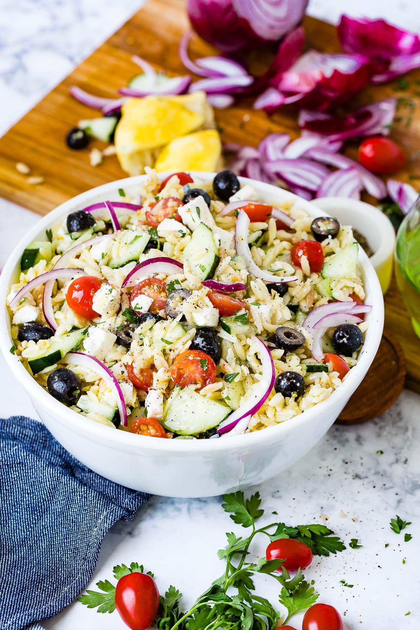 A white bowl of Greek orzo pasta salad with slices of red onion, cherry tomatoes, cucumbers and black olives.