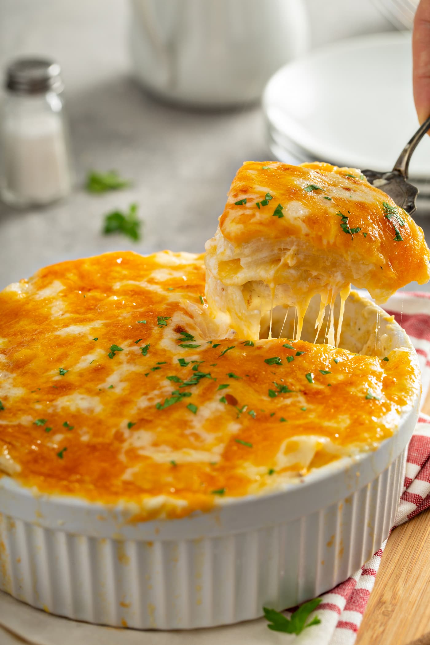 A serving of instant pot cheesy potatoes are being lifted from a white casserole dish with a serving spoon. the melted cheese is stringing out below the spoon. there is a stack of dinner plates in the background and a red and white kitchen towel beside the casserole dish.