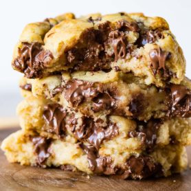 a photo of four thick melty chocolate chip cookies that have been broken in half stacked on top of each other on top of a wooden plate.