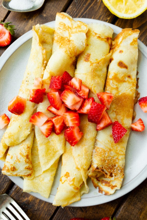 a photo of four swedish pancakes rolled up and sitting next to each other on a white plate topped with diced strawberries.