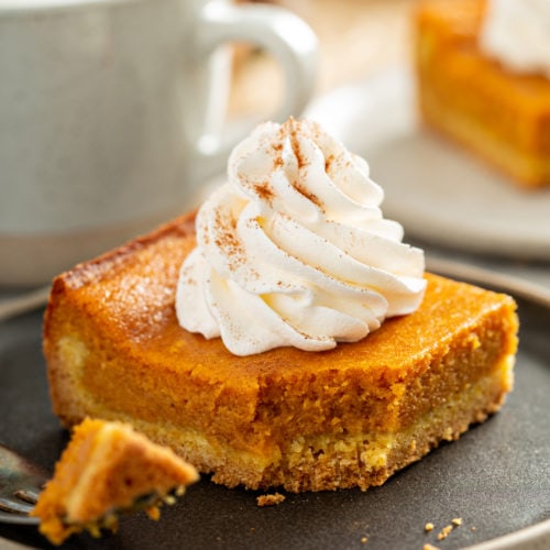 A square piece of pumpkin gooey butter cake with a swirl of whipped cream on top. A fork with a bite of cake is laying on the plate and a white cup and white plate with a piece of pumpkin cake are in the background.