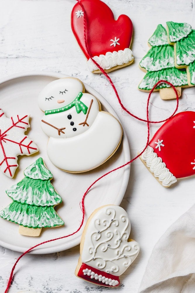 Christmas sugar cookies decorated with royal icing. There is a snowman with a green scarf, a red mitten, a snowflake with red accents, a green tree with white snow.