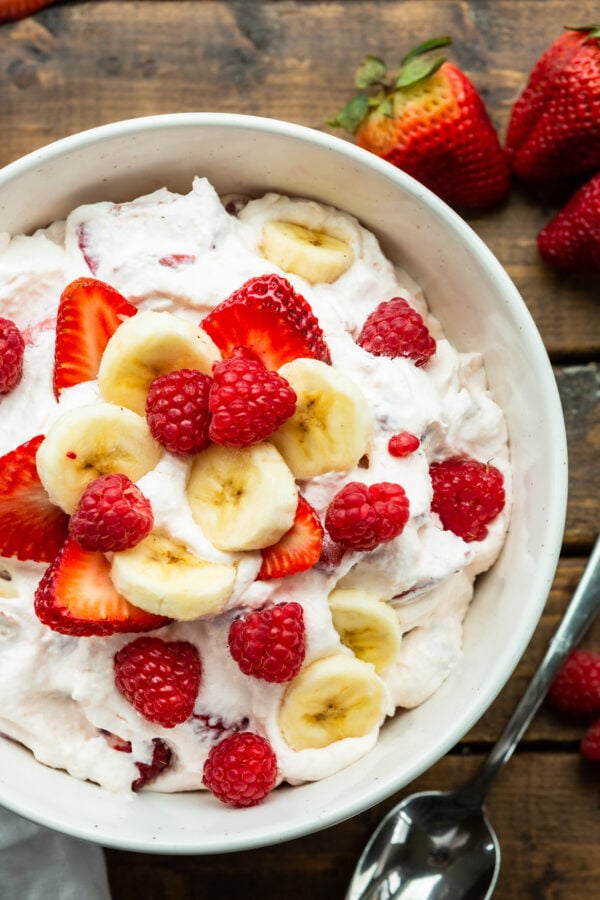 a photo of a bowl of creamy cheesecake fruit salad topped with slices of banana and whole raspberry and sliced strawberries