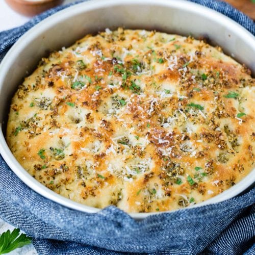 A baking dish with freshly baked Garlic Parmesan focaccia bread. It is golden brown with bits of green herbs.