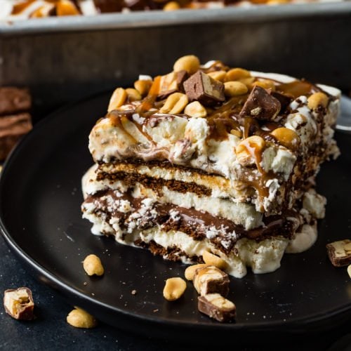 A square piece of snickers ice cream cake on a brown dessert plate. There are layers of ice cream, chocolate cake, fudge, caramel and peanuts. There is fudge and whipped cream on top and salted peanuts are sprinkled over the whole thing. A pan of snickers ice cream cake is in the background.