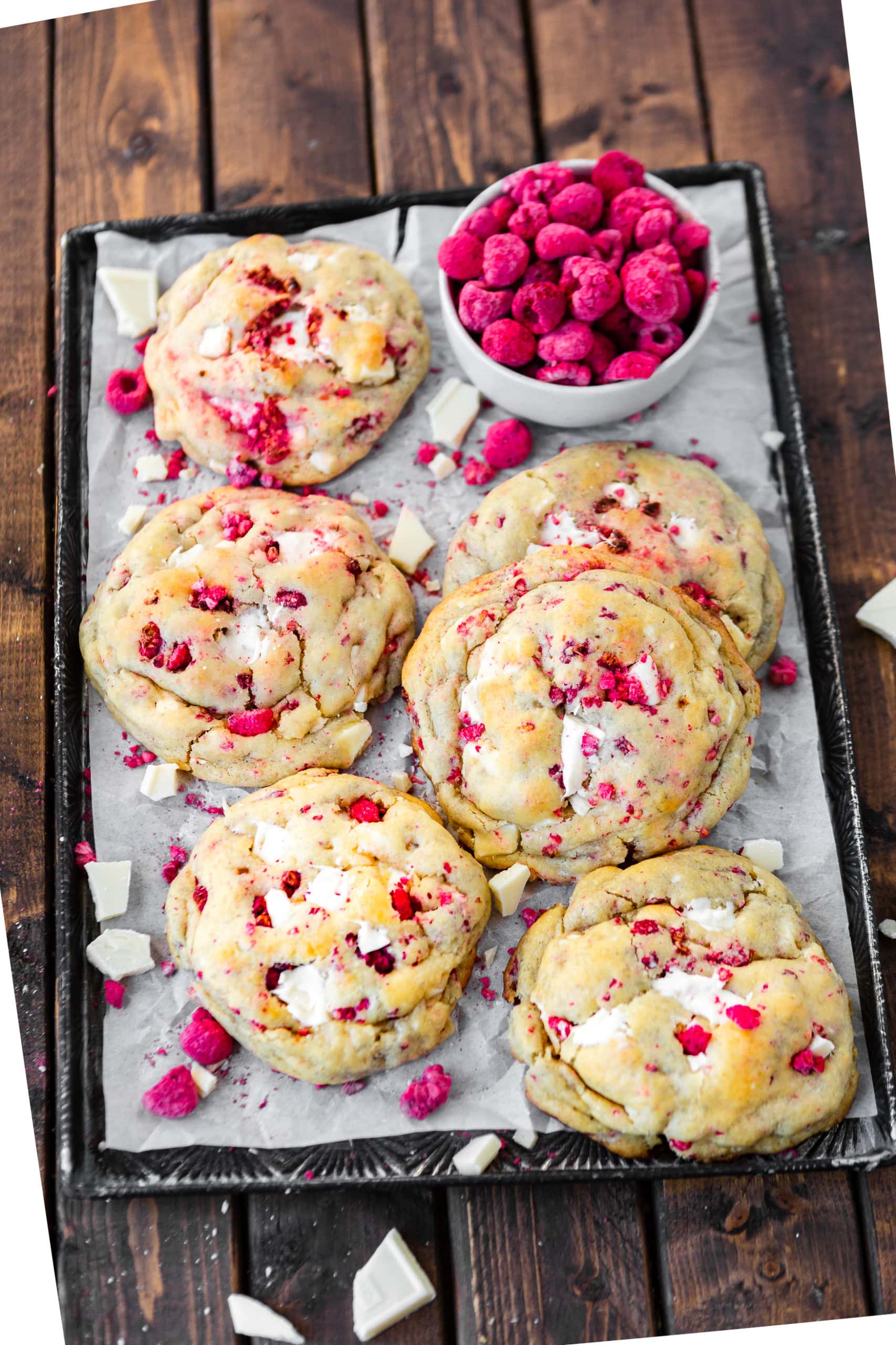 A photo of a baking sheet lined with white parchment paper and topped with size large golden baked white chocolate raspberry cokies with a small bowl of whole freeze dried raspberries and chunks of white chocolate and pieces of freeze dried raspberries scattered around the cookies and baking sheet.