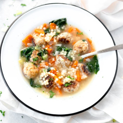 A bowl of Italian wedding soup. A spoon is in the soup and it is packed with meatballs, carrots and spinach.