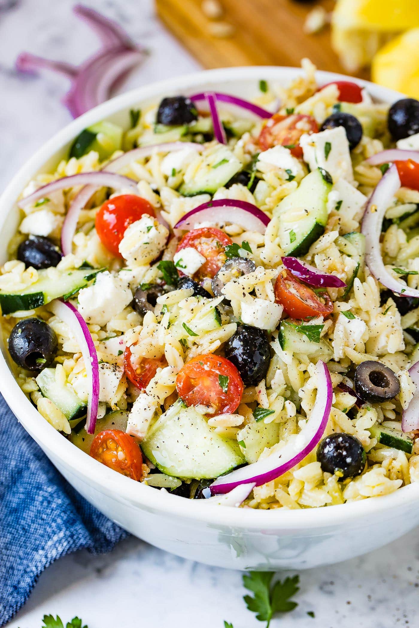 A white bowl of Greek orzo pasta salad with halved cherry tomatoes, slices of red onion, cucumber, black olives and feta cheese.