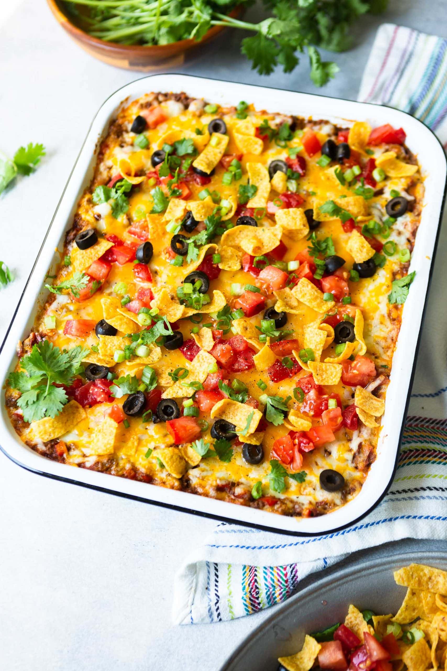 a photo of a casserole dish full of taco casserole topped with diced tomatoes, fritos chips, sliced black olives, chopped cilantro and diced green onion.