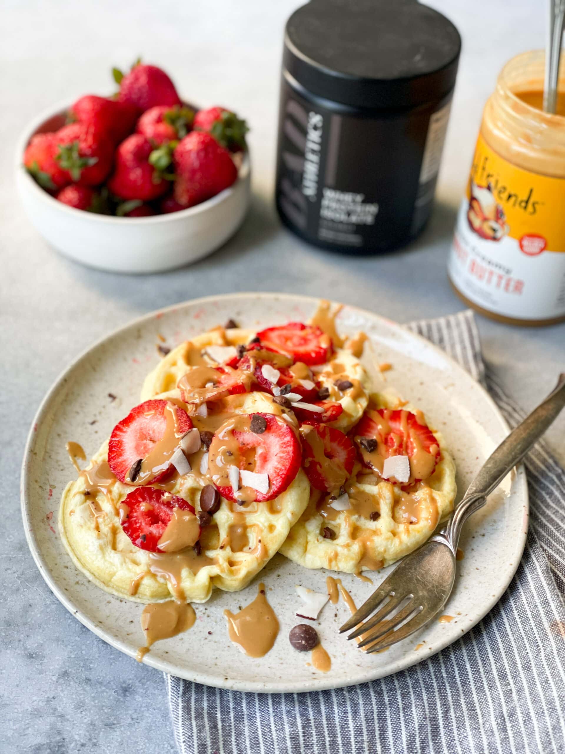 a photo with three small circular waffles sitting on a white dinner plate topped with sliced strawberries, chocolate chips and peanut butter drizzle. There is a bowl of whole strawberries sitting in the background sitting next to a black container of protein powder and jar of peanut butter.