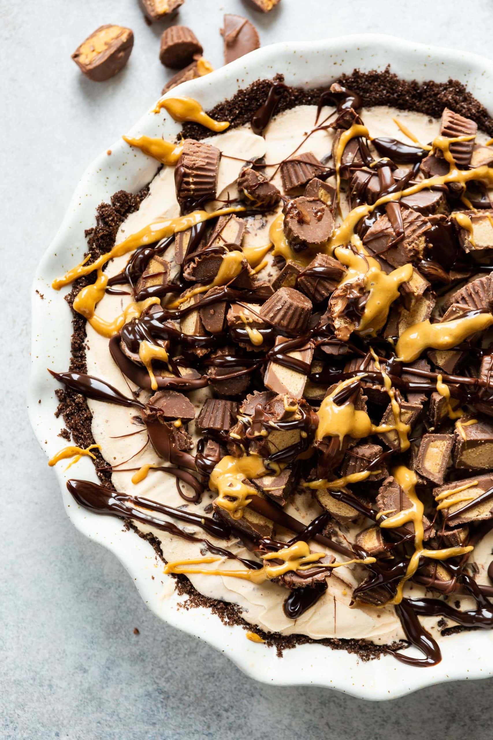 A photo of a reeses ice cream pie in a white pie dish topped with reeses peanut butter cups and chocolate and peanut butter drizzle.